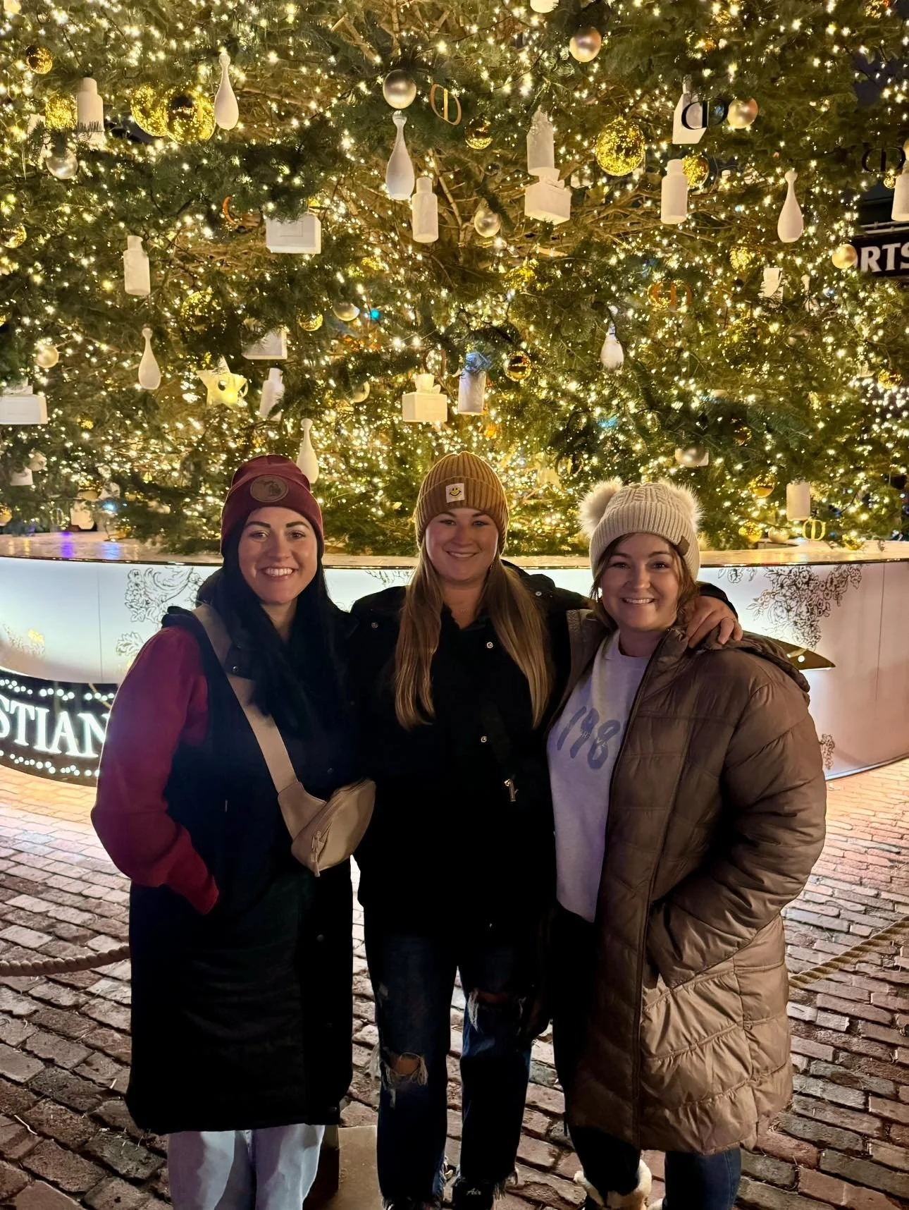 Three women standing together in front of a large, decorated Christmas tree at night, smiling, with lights and ornaments.