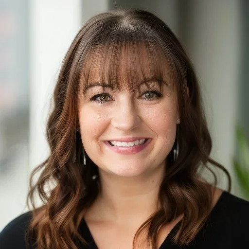 A smiling woman with shoulder-length brown hair, wearing a black top, standing indoors with blurred windows and plants in the background.