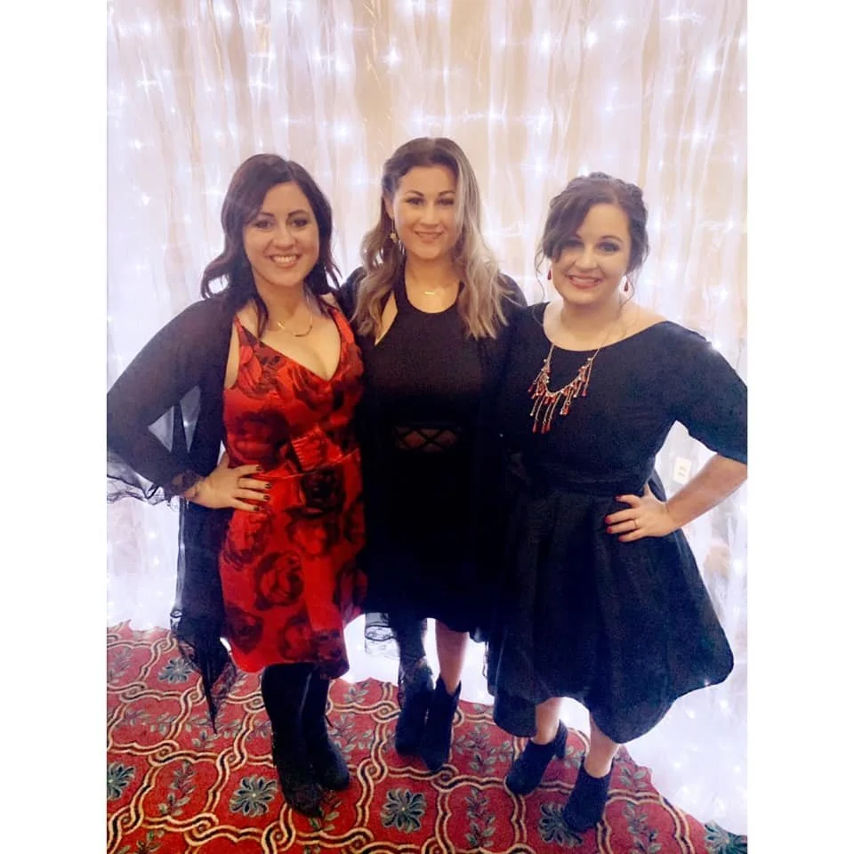 Three women dressed in formal attire standing together, smiling for a photo in front of a light-colored, illuminated curtain.