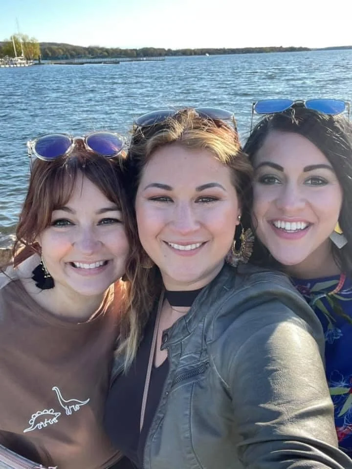 Three women smiling taking a selfie by a lake, with trees and a pier in the background.