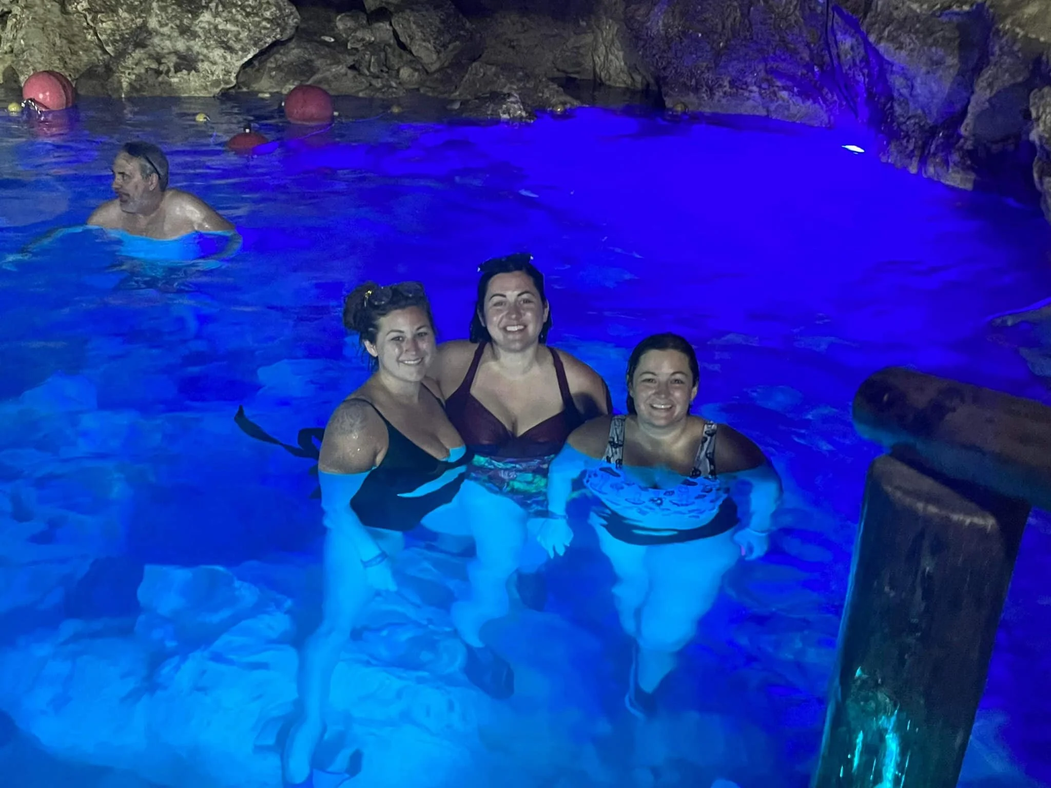 Three women and one man enjoying a swim in a cave-like pool with blue lighting.
