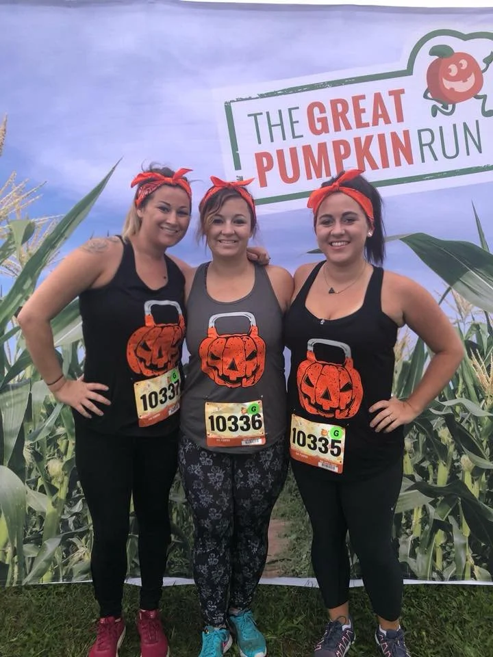 Three women in athletic wear with orange bandanas on their heads, standing outdoors in front of a backdrop with a cornfield and a sign that reads 'The Great Pumpkin Run', all smiling and posing for a photo.