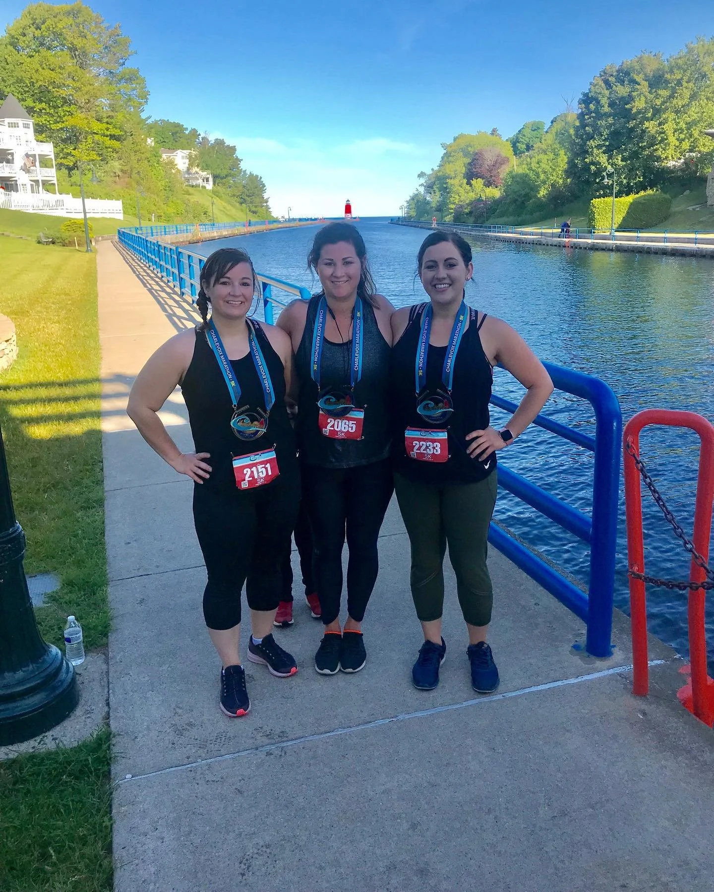 Three women in athletic attire smiling and standing together by a canal with a red lighthouse in the distance, wearing medals and race bibs.