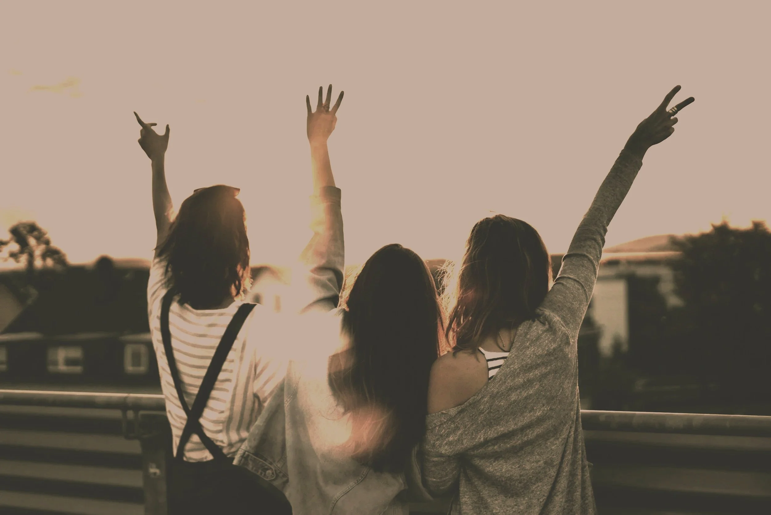Three young women sitting on a railing at sunset with their backs to the camera, raising their hands and making peace signs.