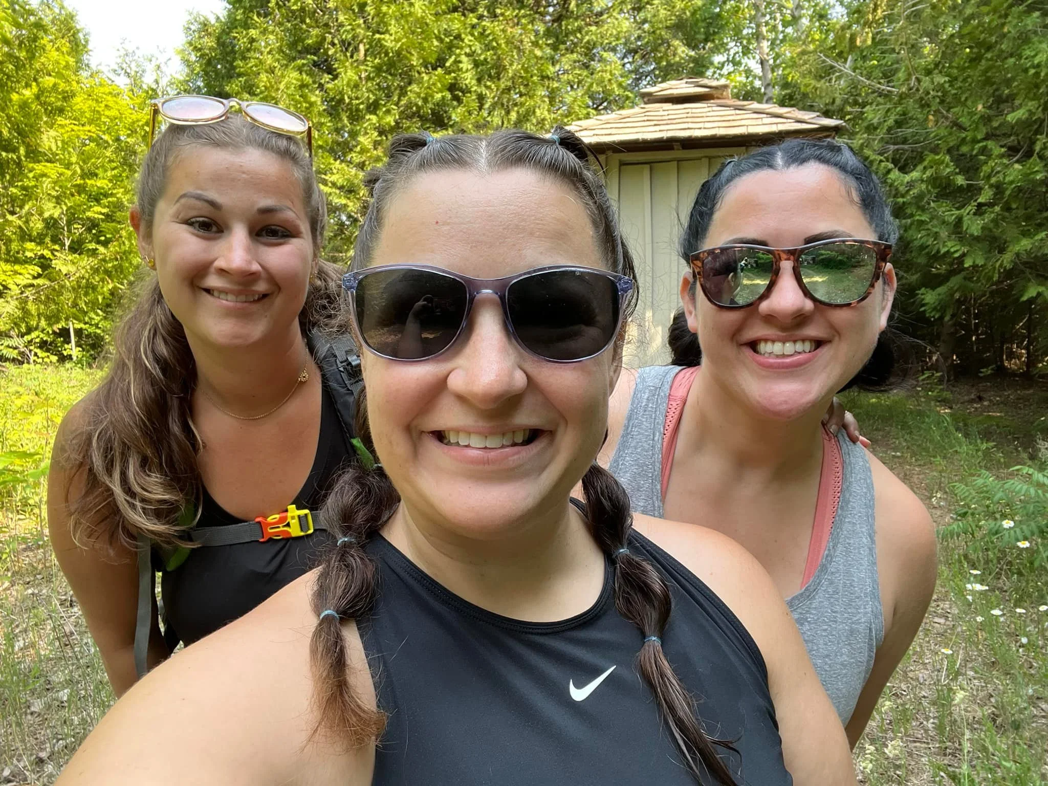 Three smiling women taking a selfie outdoors in a forested area with trees and a small yellow shed in the background.
