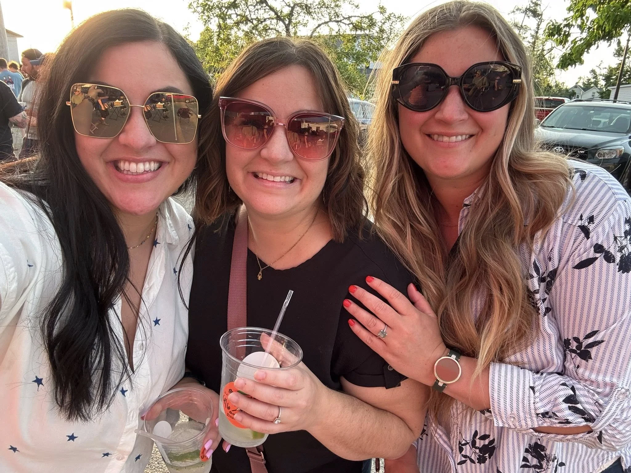 Three women smiling wearing sunglasses, standing outdoors in a social setting, dressed casually, with cars and trees in the background.
