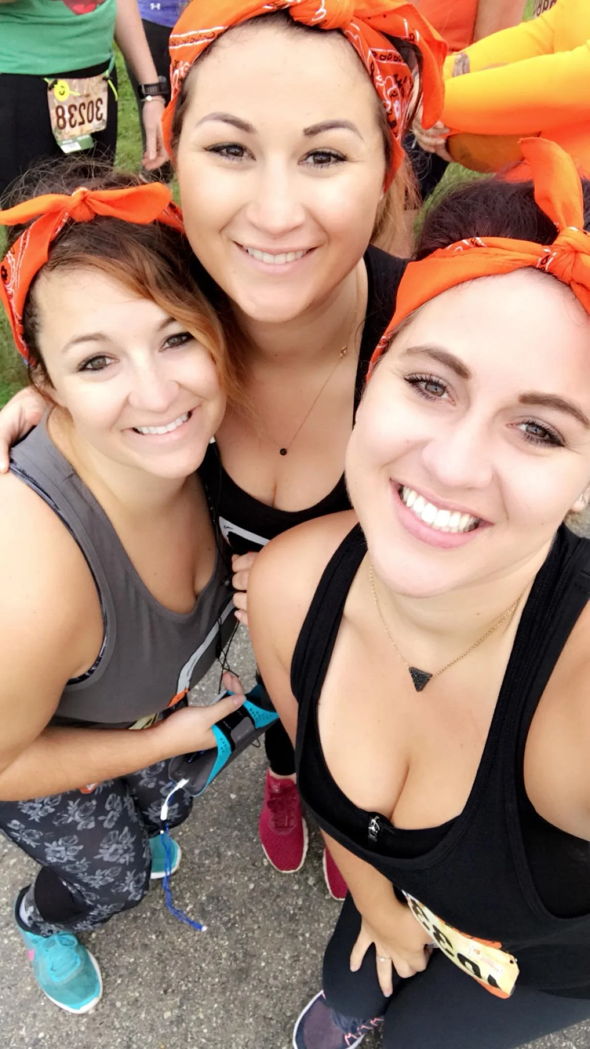 Three women wearing orange bandanas on their heads, smiling for a selfie during a group outdoor running event, with two wearing athletic black tops and one in a gray tank top.