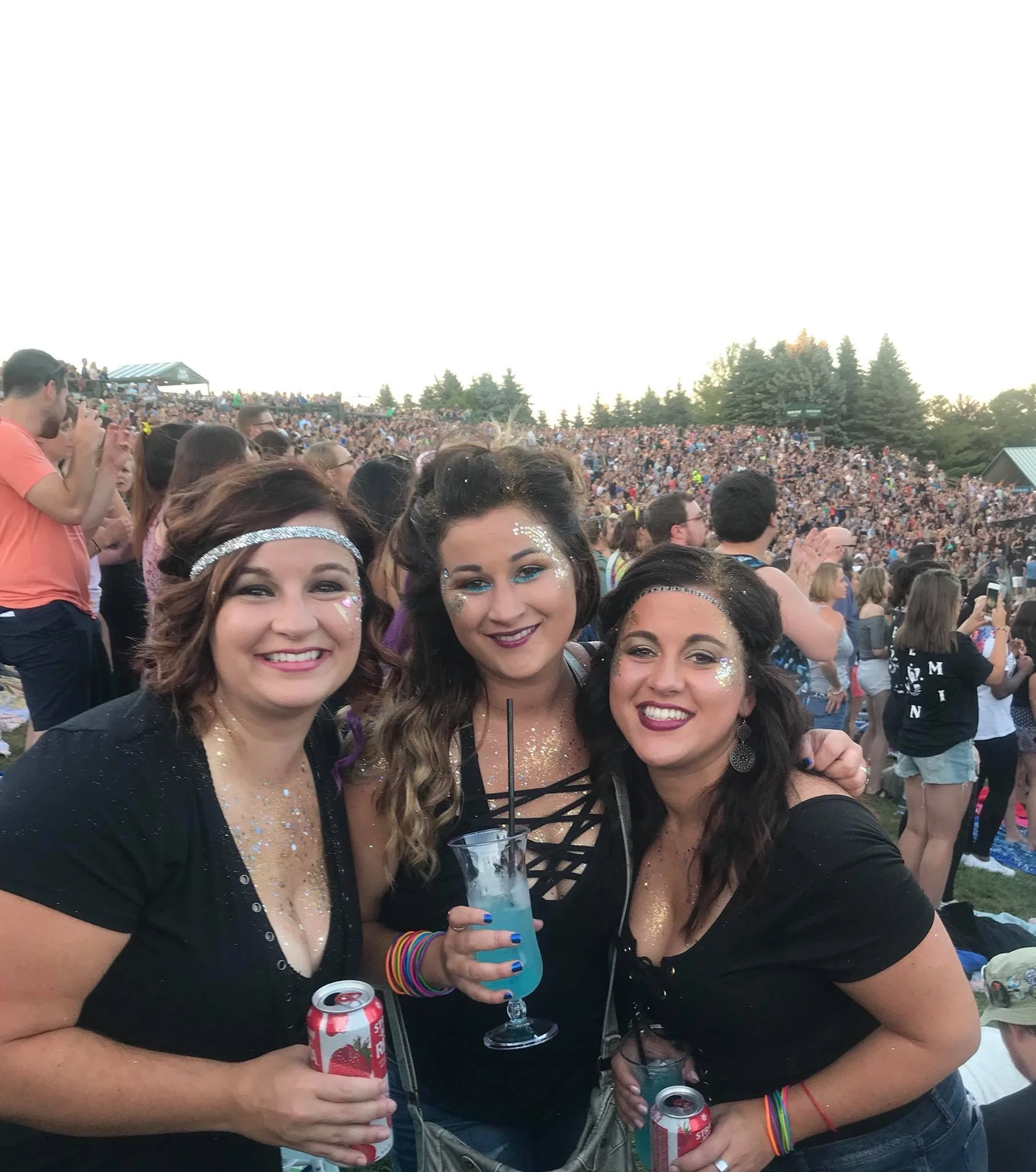 Three women at an outdoor concert or festival, holding drinks and smiling, with a large crowd in the background.