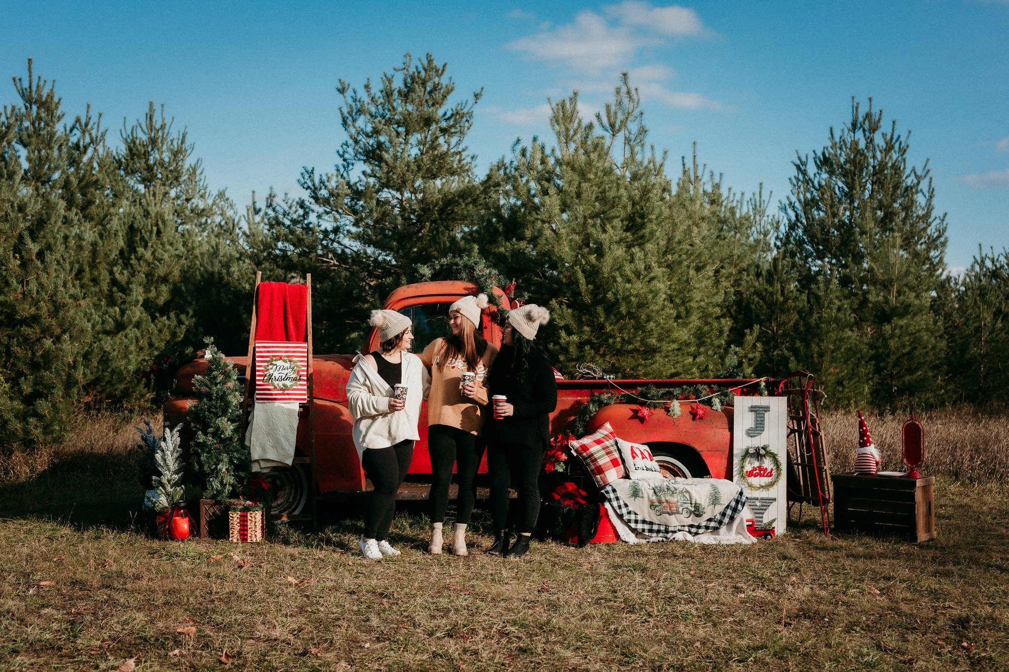 Three women in winter hats and casual clothes standing in front of a vintage red truck decorated for Christmas with holiday decorations, a small Christmas tree, and presents in an outdoor setting with evergreen trees in the background.