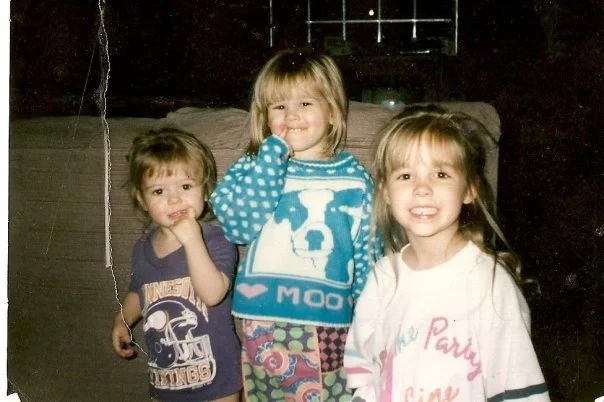 Three young girls smiling and posing together indoors, one wearing a patterned sweater, another in a white shirt, and the third in a dark t-shirt with a graphic design.