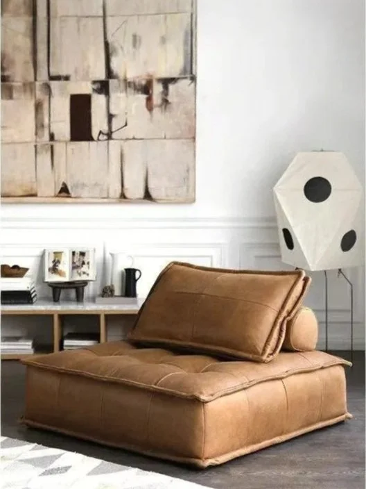 A tan, low-profile sofa with a tilted backrest in a modern living room. Behind it, a white sideboard with framed pictures, books, and decorative objects. To the right, a unique white paper lantern-style floor lamp with black circular accents. A large abstract painting hangs on the wall above the sideboard.