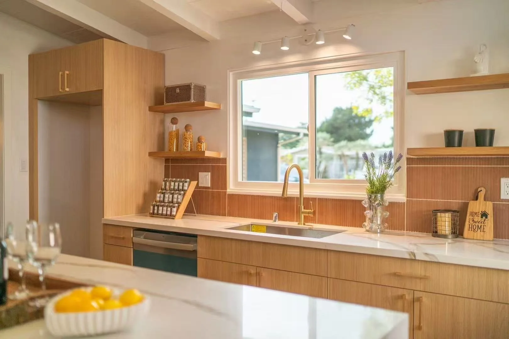 A modern kitchen with wooden cabinets, a white marble countertop, a gold faucet, and a large window overlooking a backyard with trees.