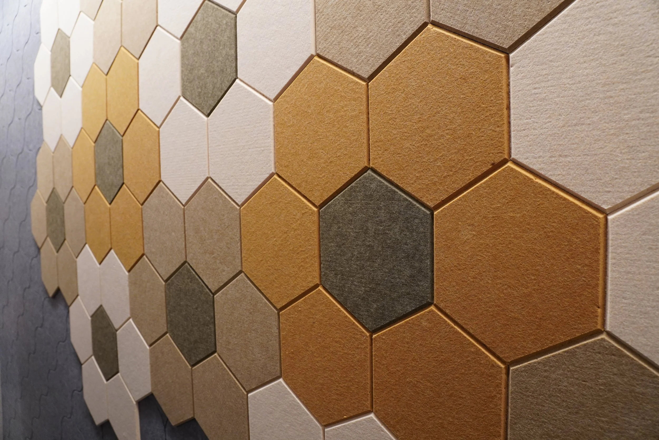 Close-up of various hexagonal tiles in shades of beige, brown, gold, and gray, arranged in a honeycomb pattern on a wall.