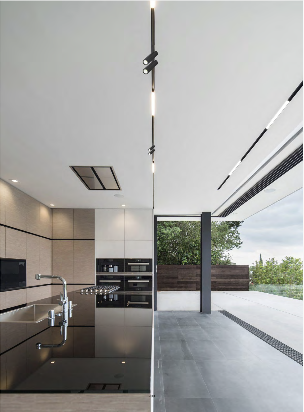 Modern kitchen with brown cabinetry, built-in black appliances, a stainless steel sink, and a large window overlooking greenery and outdoor patio.