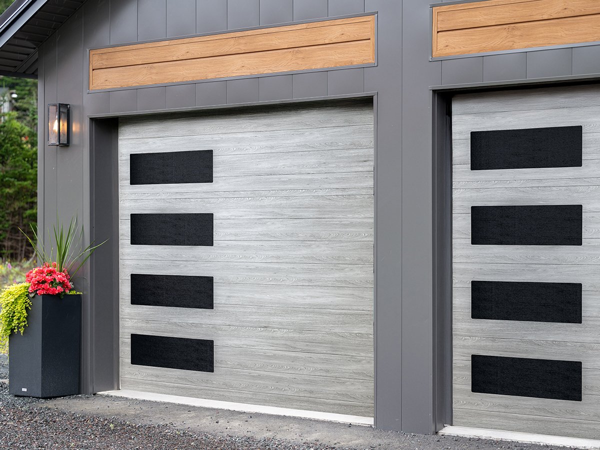 Modern garage door with horizontal wooden panels, grey base, and black rectangular window accents. A potted plant with pink flowers is on the left side.