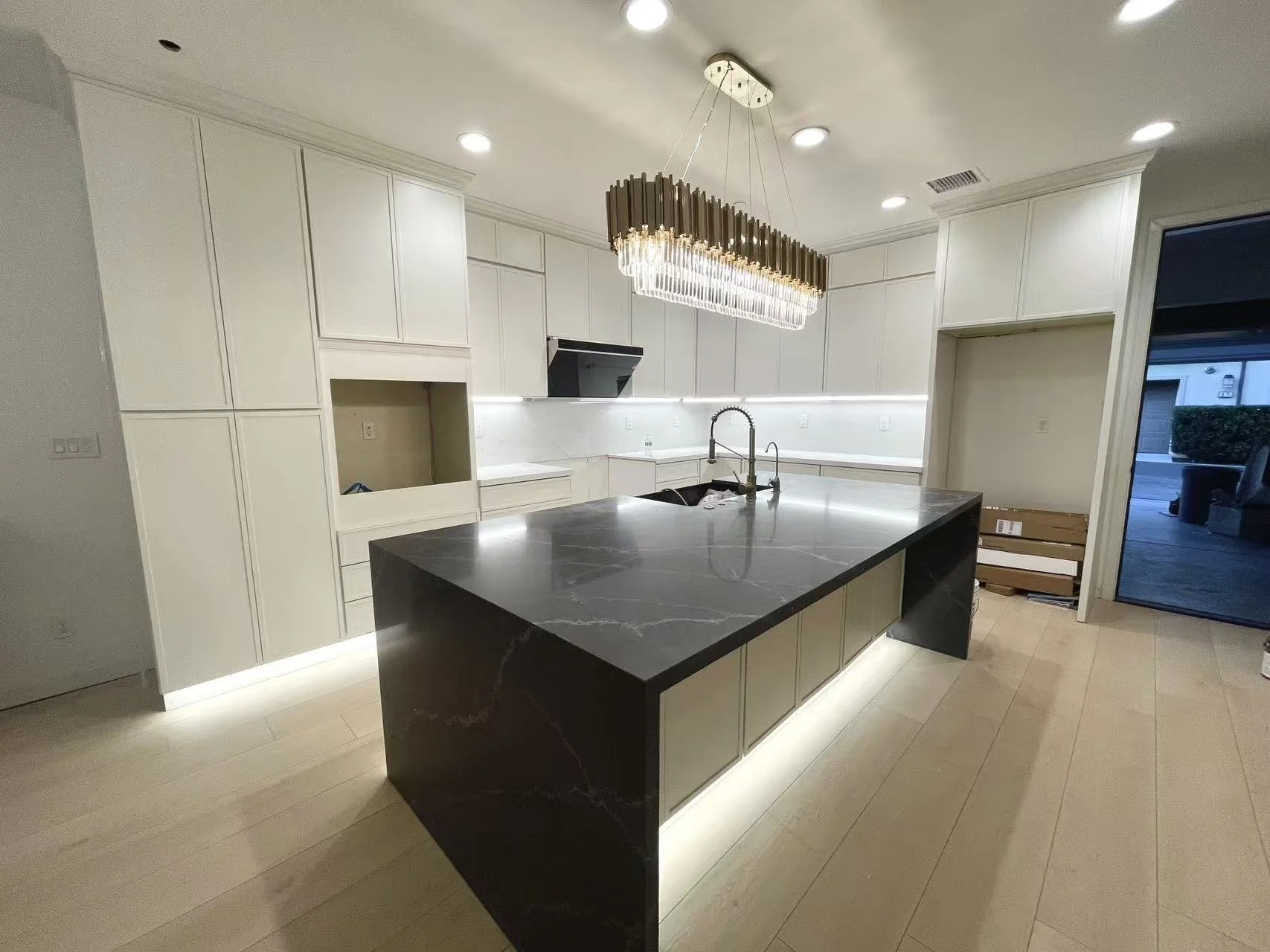 Modern kitchen featuring white cabinetry, a black marble island with under-cabinet lighting, a chandelier above, and a sliding glass door leading outside.