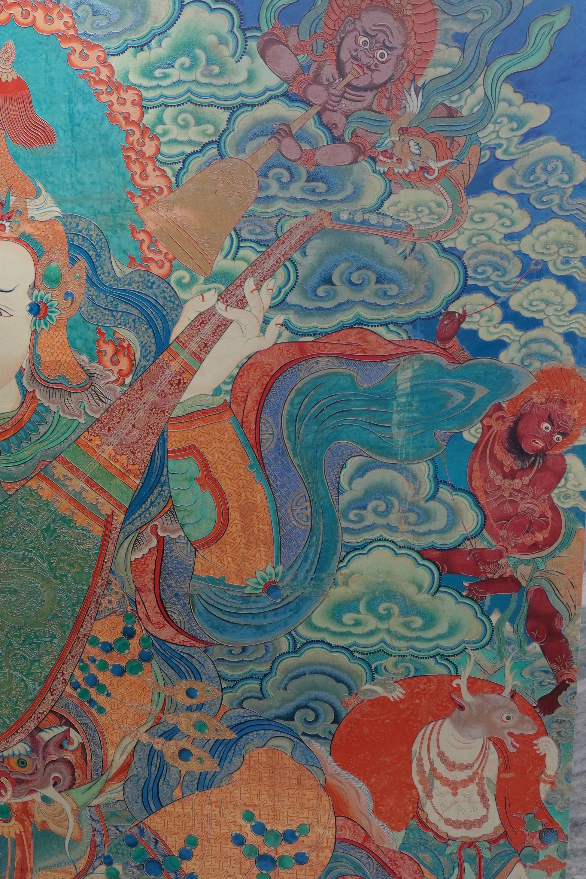 Colorful Tibetan religious painting showing a deity playing a flute, surrounded by clouds, mythical creatures, and a bull, with intricate patterns and vibrant colors.