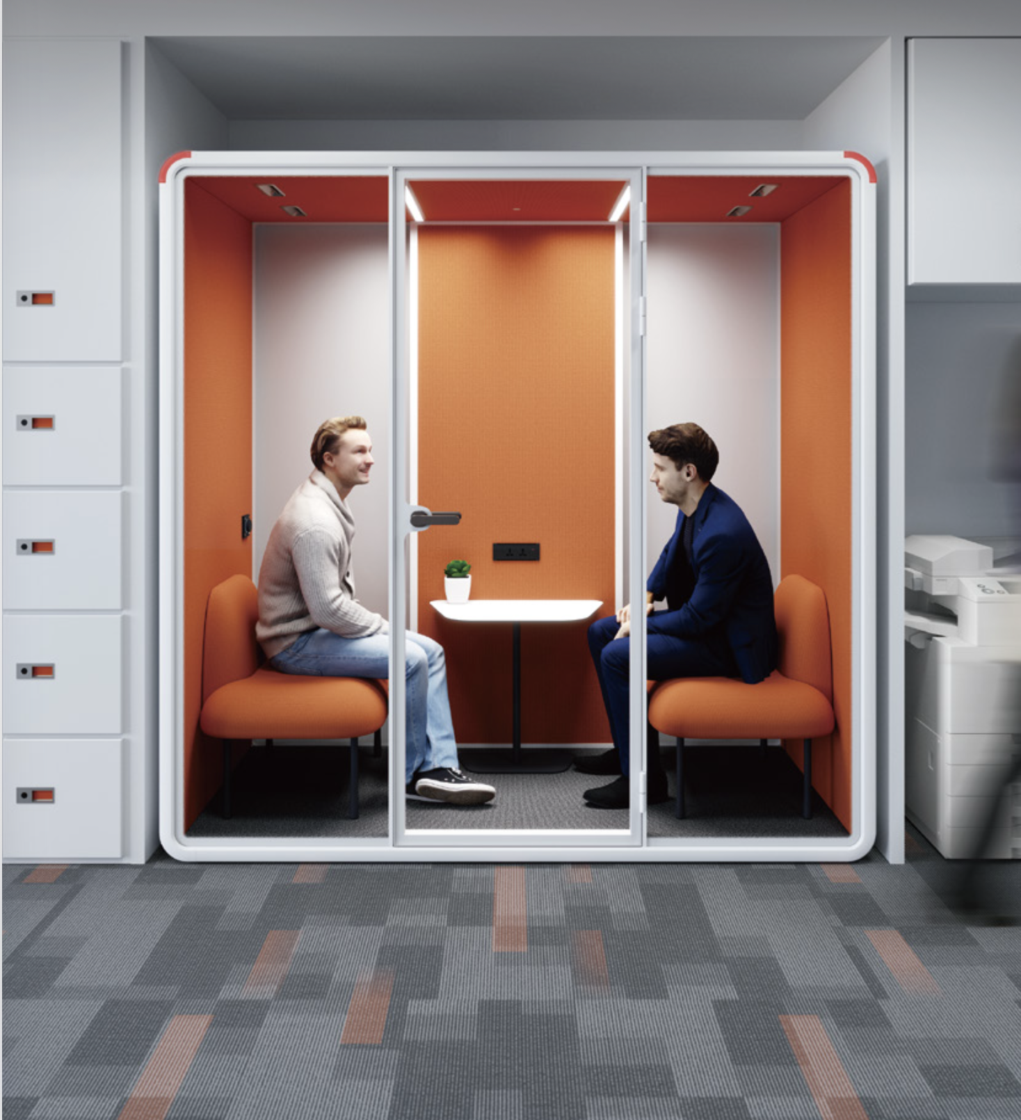 Two people sit in a modern, orange-walled phone booth in an office, engaged in conversation. The booth has glass doors, orange cushioned seats, a small white table with a plant, and is located next to lockers and office equipment.