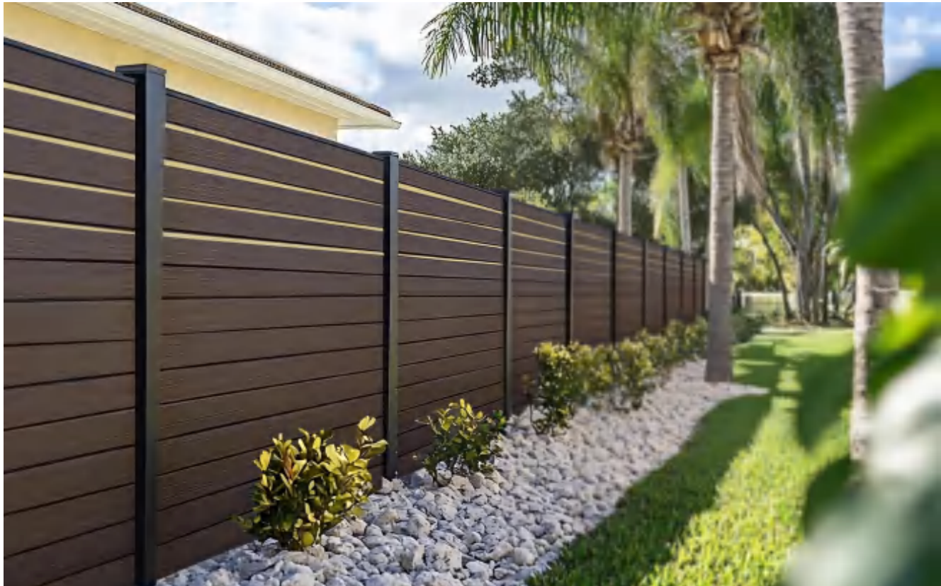 A backyard with a dark brown wooden fence and yellow and green shrubs, with palm trees in the background and a grassy lawn.