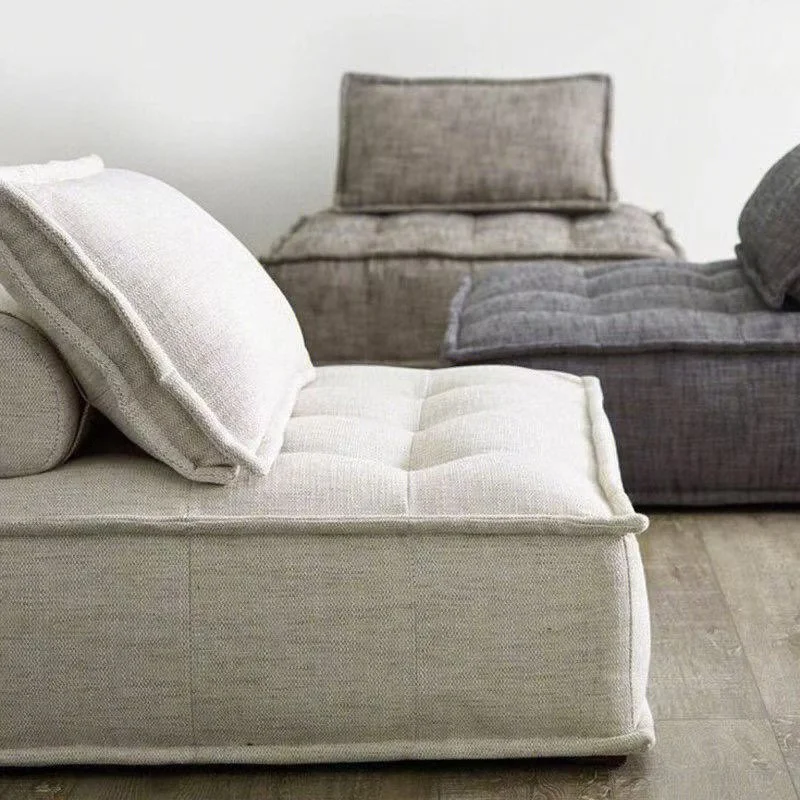 A beige upholstered sofa with matching cushions in the foreground and a gray sofa with cushions in the background, in a minimalistic room with a wooden floor and white wall.
