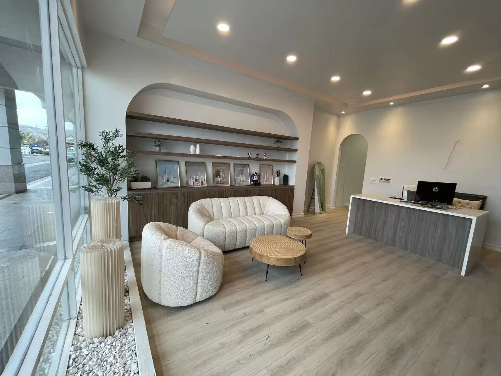 Modern hotel lobby with beige and wooden furniture, a reception desk, a seating area with a sofa and armchair, large windows, and decorative shelves with framed pictures and plants.