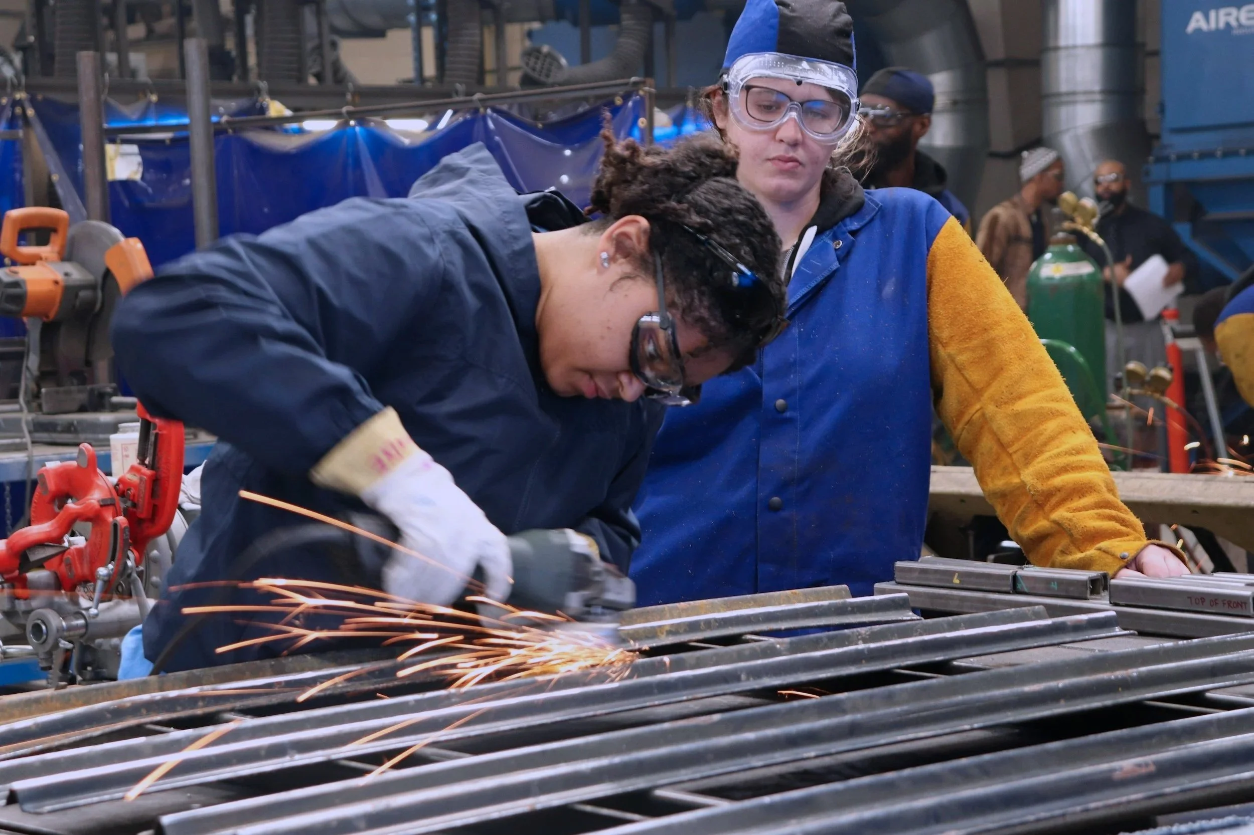 Two women working with metal in a workshop, one using a power tool that sparks, the other observing. Both wearing safety glasses and protective clothing.