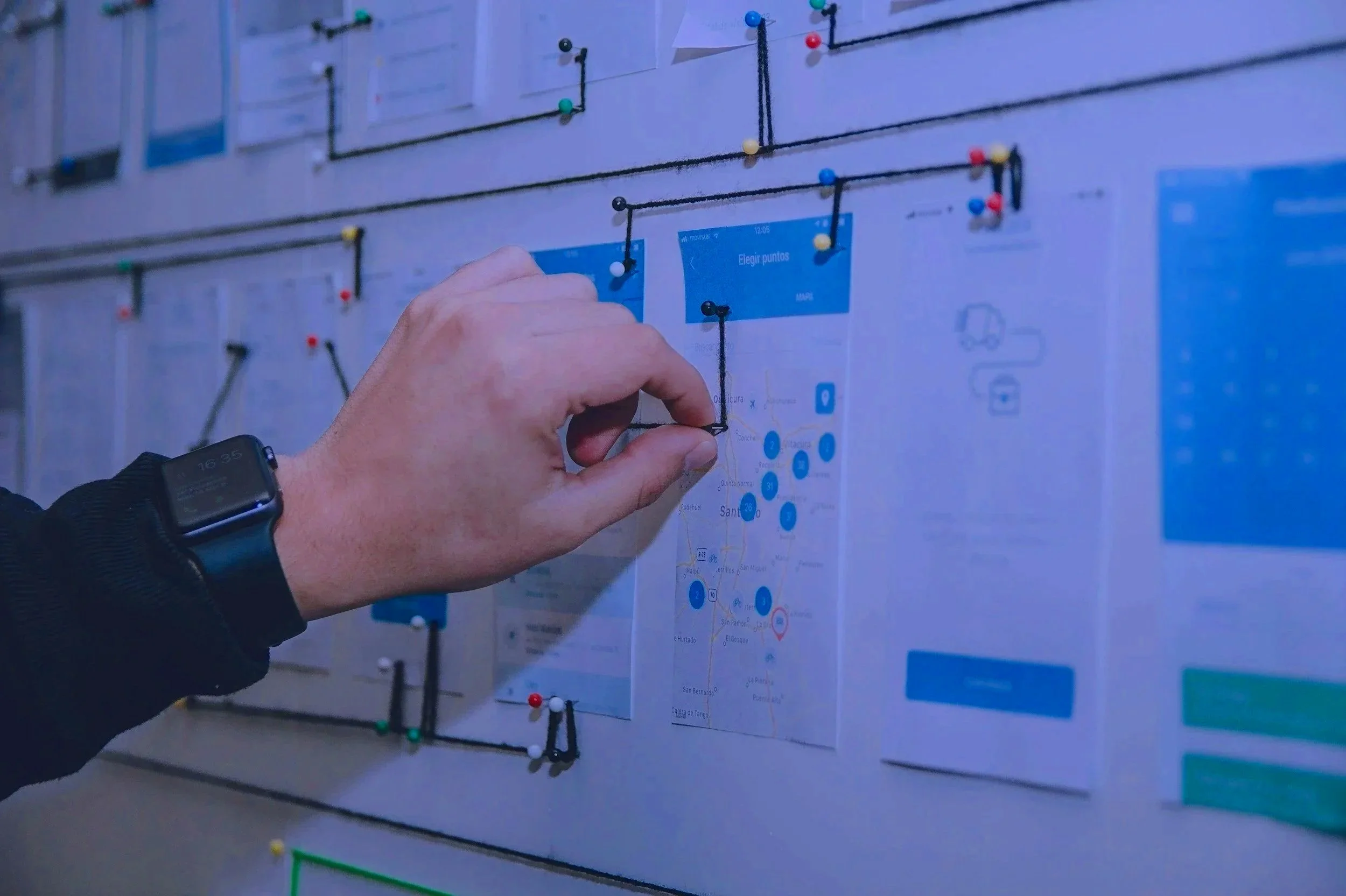 Person pinning a paper map to a wall covered with charts and notes using push pins, with a smartwatch on their wrist.