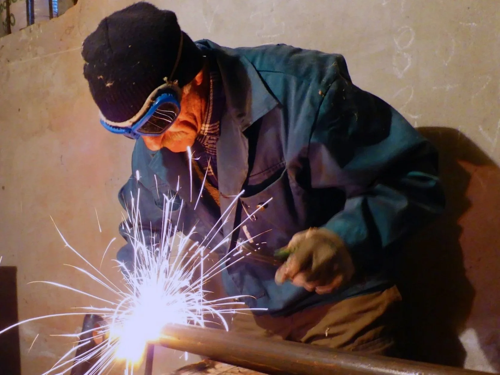 A person wearing a black hat, blue goggles, a dark green jacket, and gloves is working with a welding tool, creating bright sparks against a plain wall.