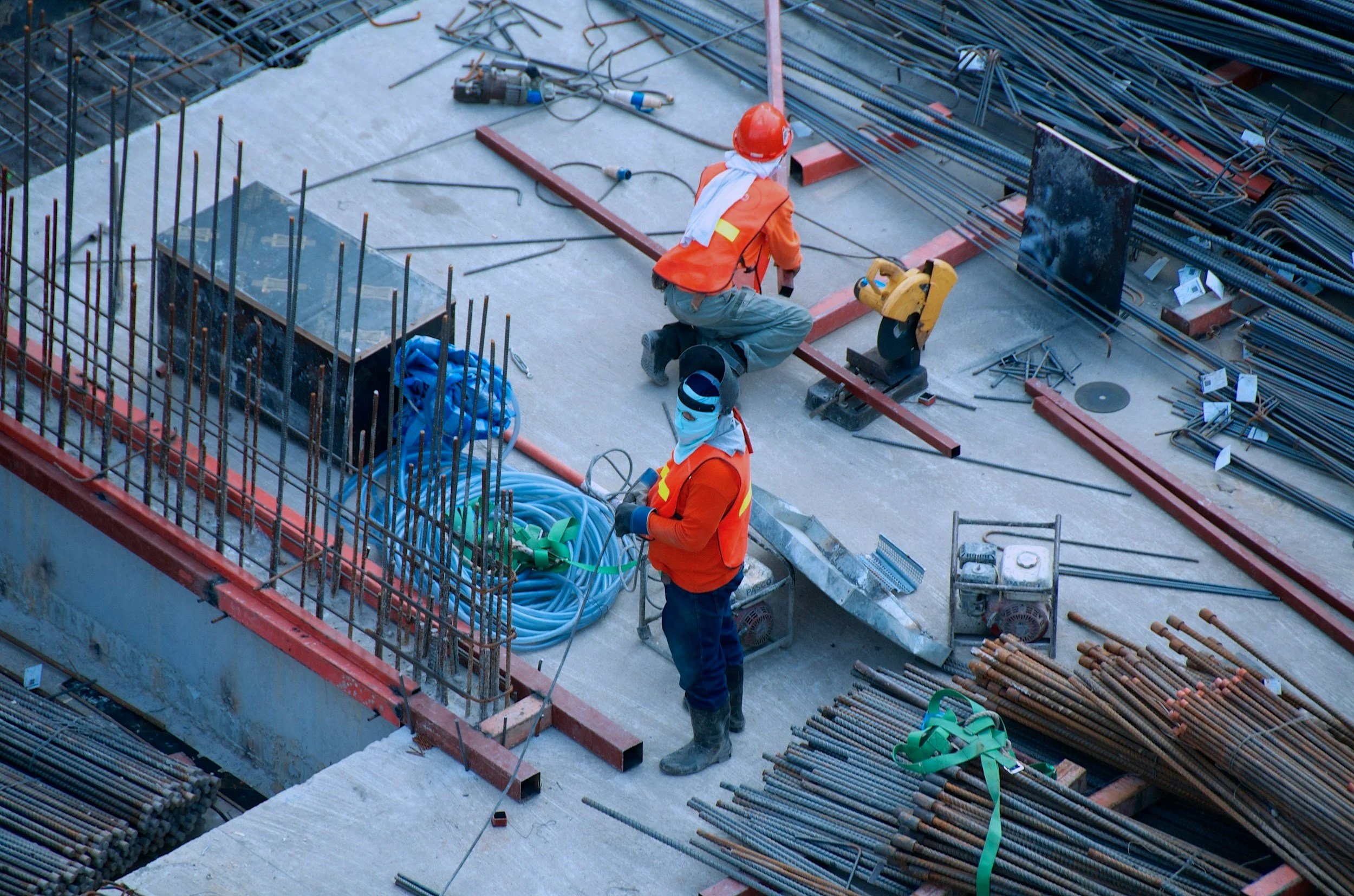 Two construction workers wearing orange safety vests and helmets working on a building site. One is kneeling while the other stands among construction materials and tools.