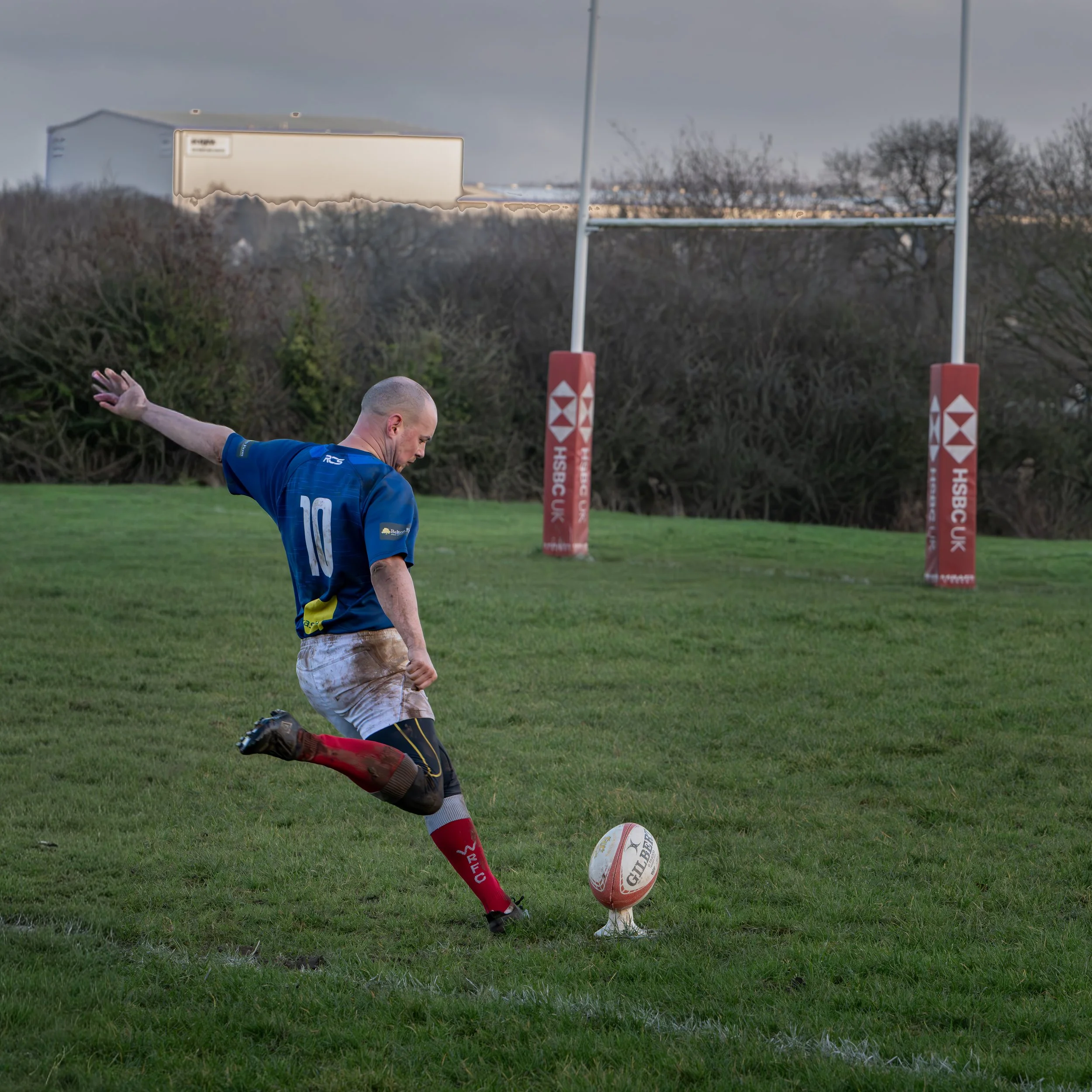A rugby player in a blue jersey with the number 10, kicking a rugby ball on a grass field near goalposts, with a cloudy sky overhead.