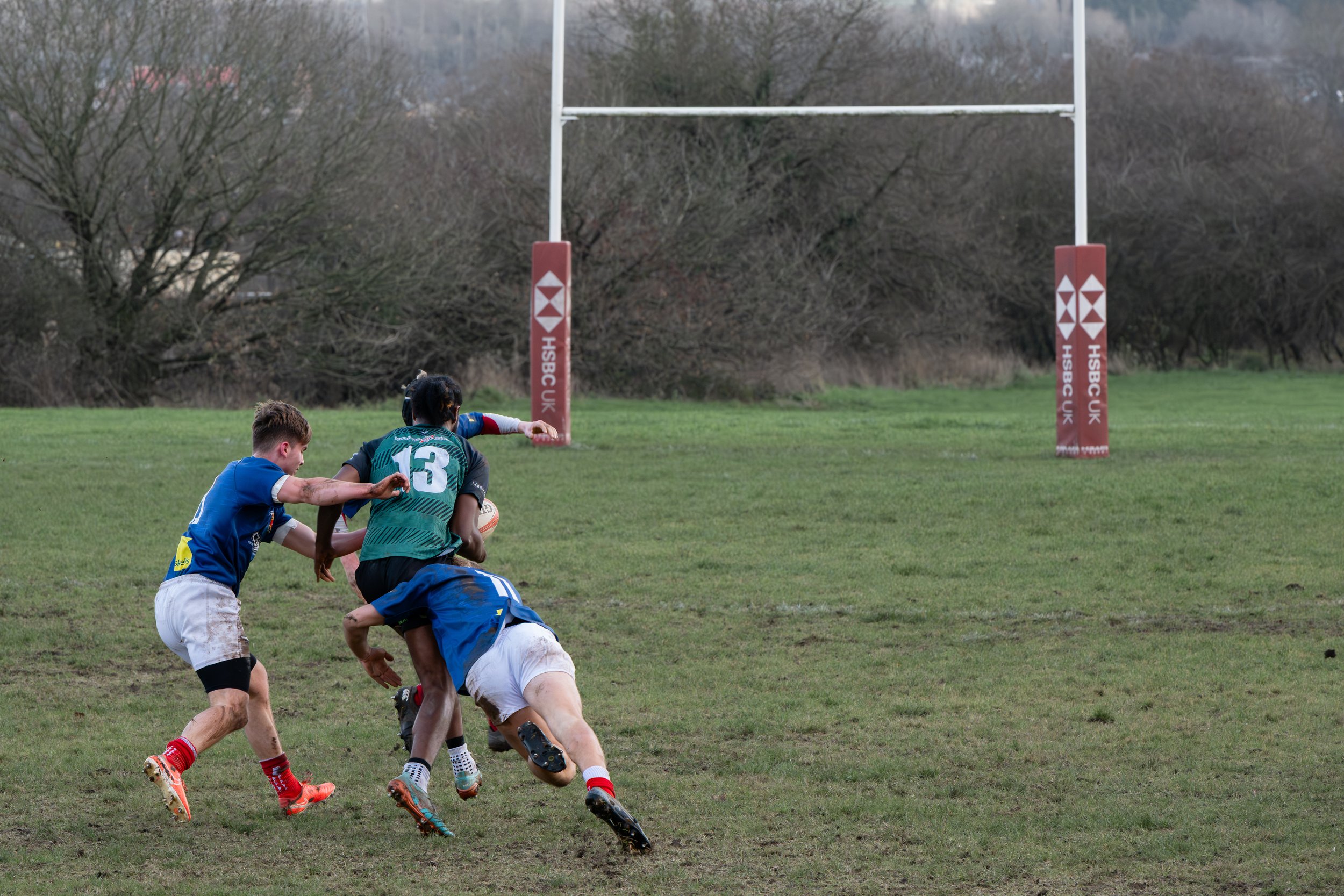 Rugby match with players contesting for the ball near an upright goalpost on a grassy field.