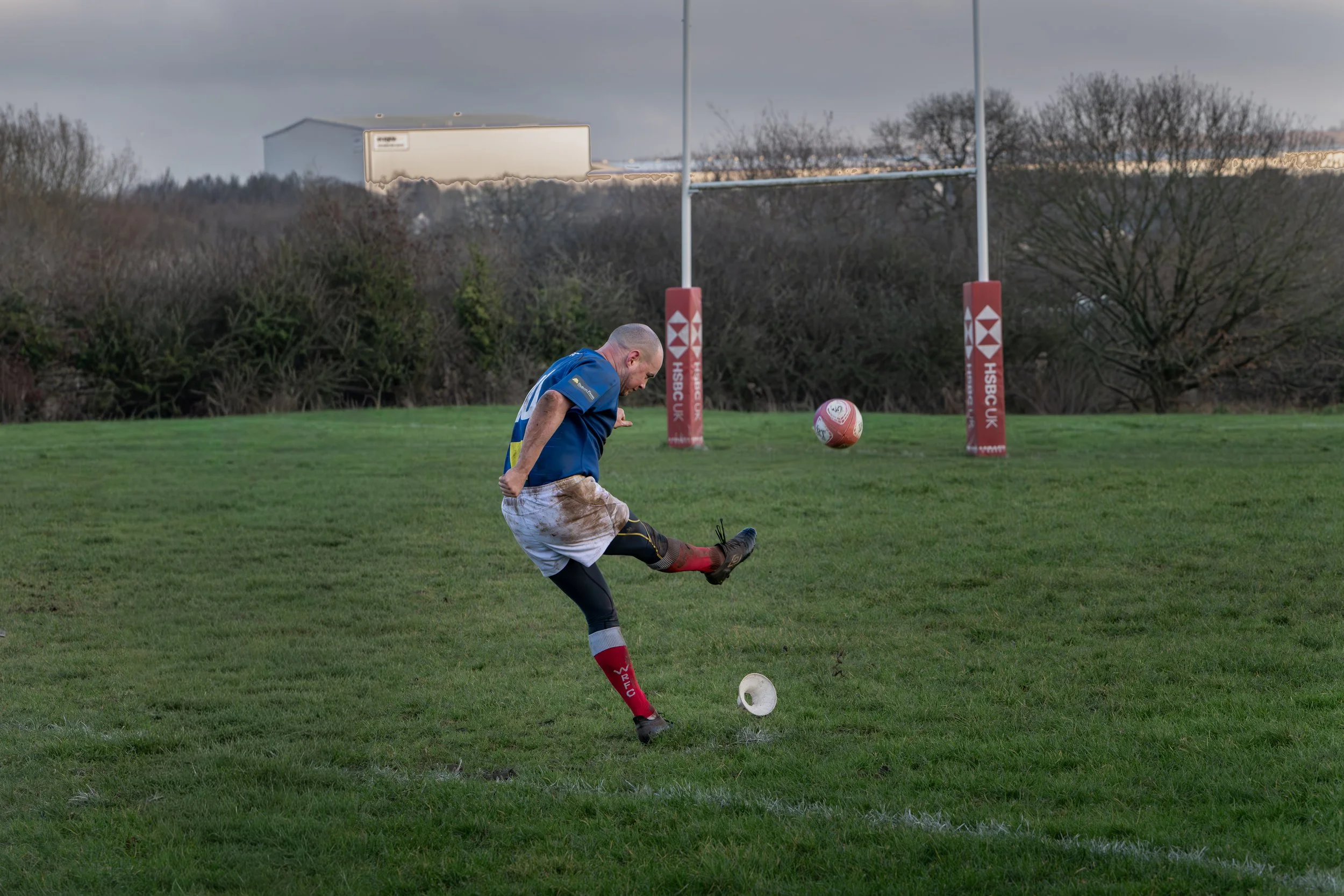 A rugby player in a blue jersey and muddy white shorts is kicking a rugby ball on a grassy field, with goalposts and trees in the background.