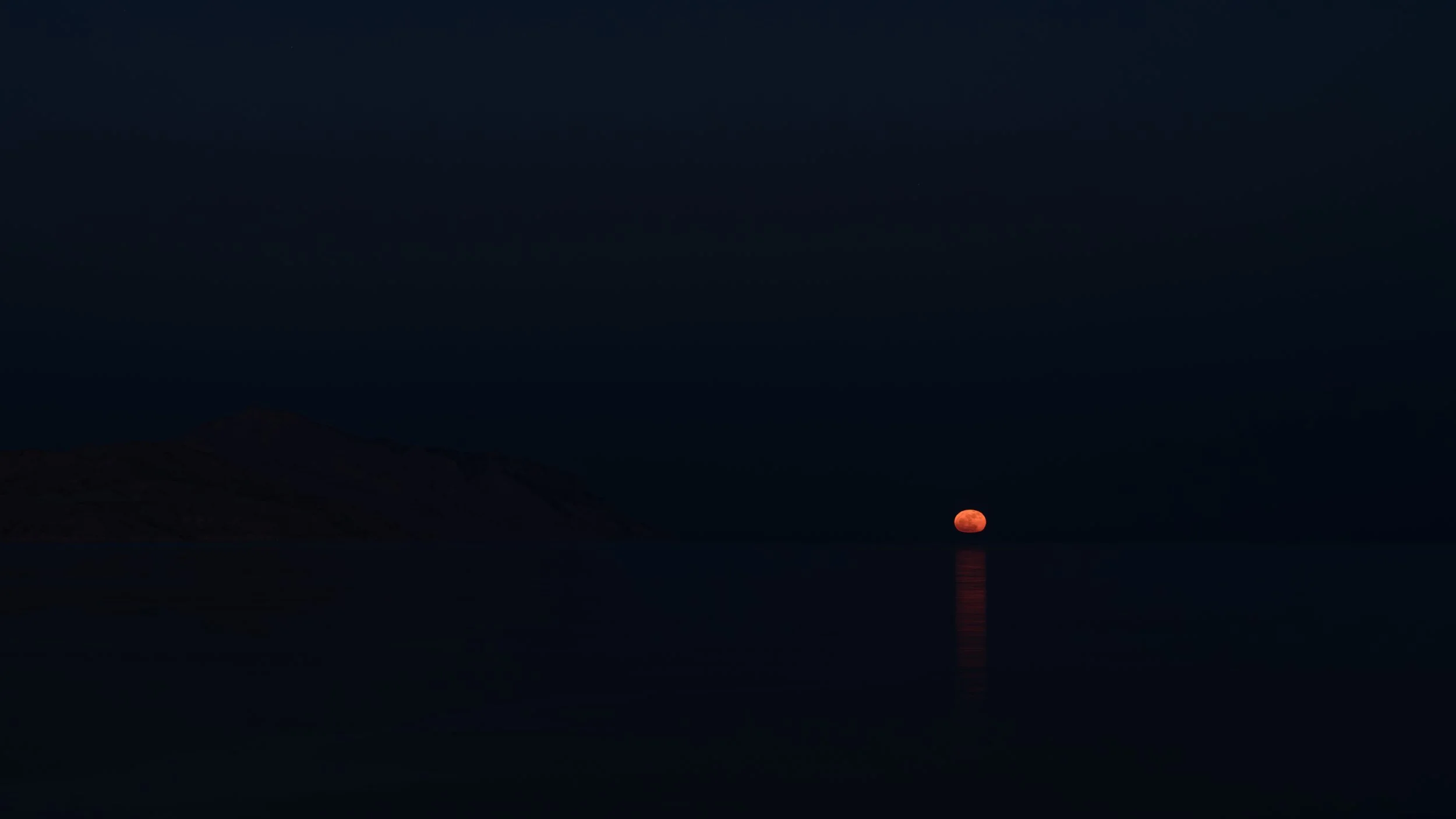 A large reddish full moon rising over a dark horizon with mountain silhouettes and a body of water reflecting the moonlight.