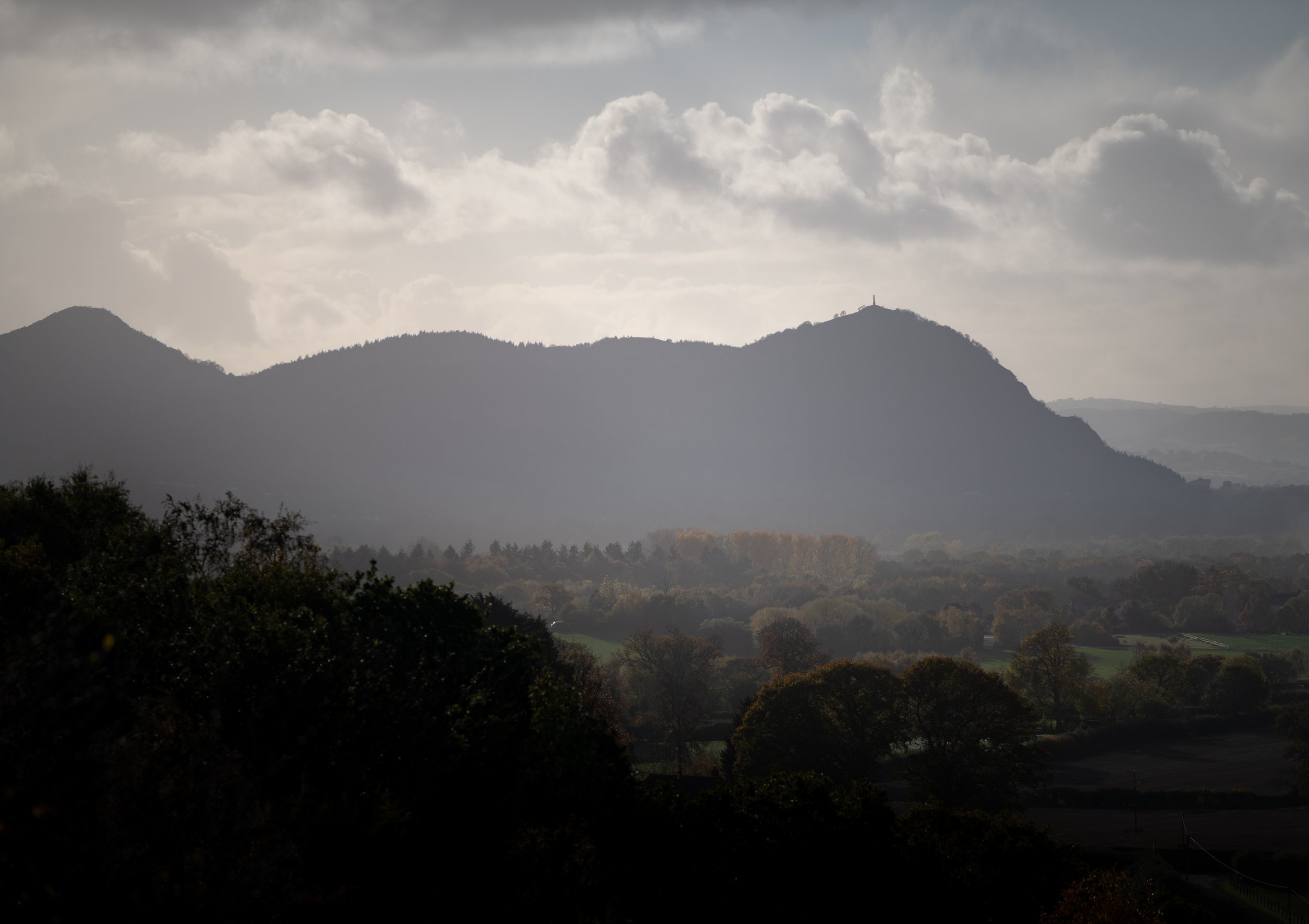 Mountains with cloudy sky and a rural landscape of trees and fields in the foreground.