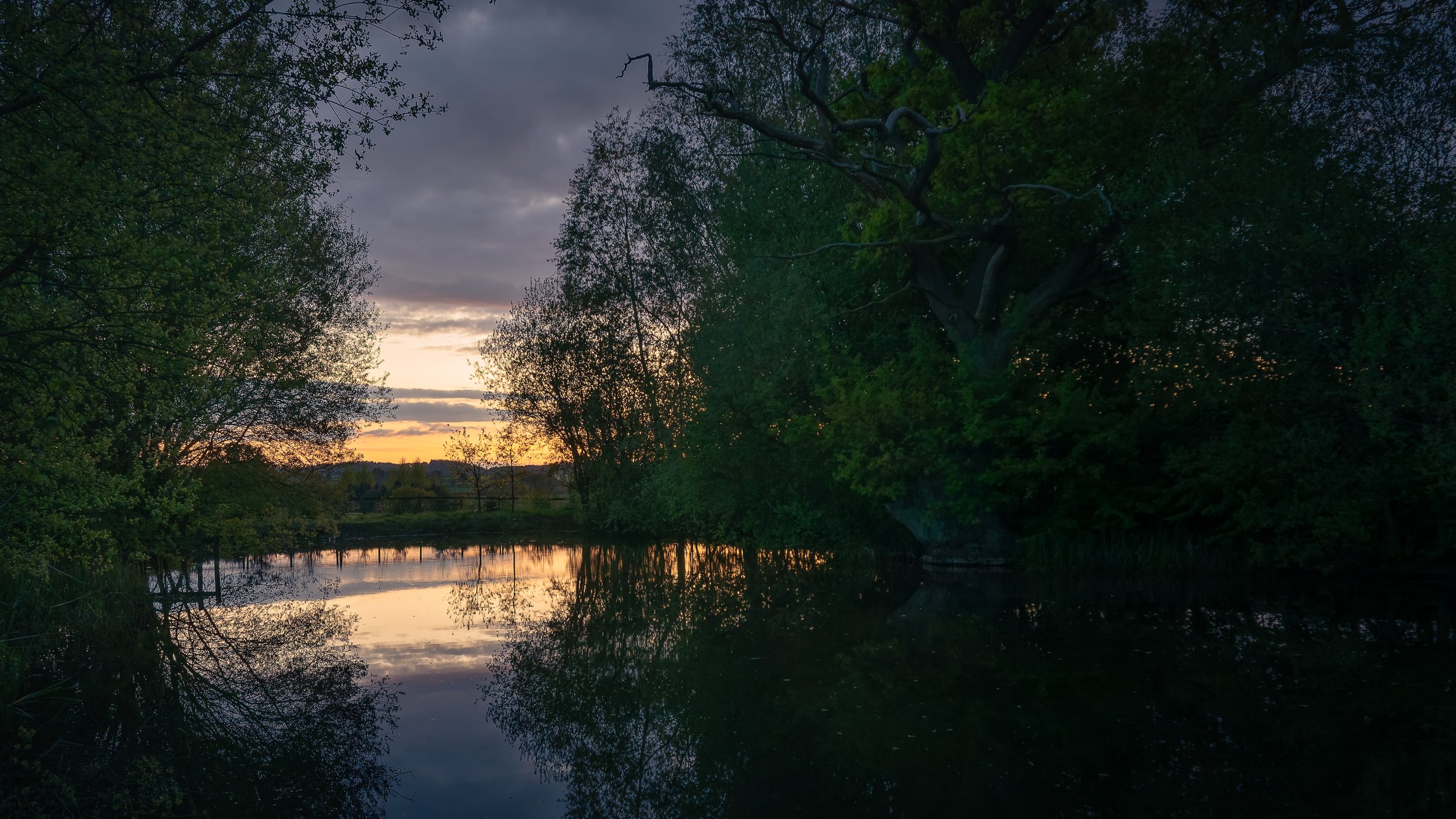 A peaceful river scene at dusk with reflections of trees and a setting sun, darkening sky with clouds.