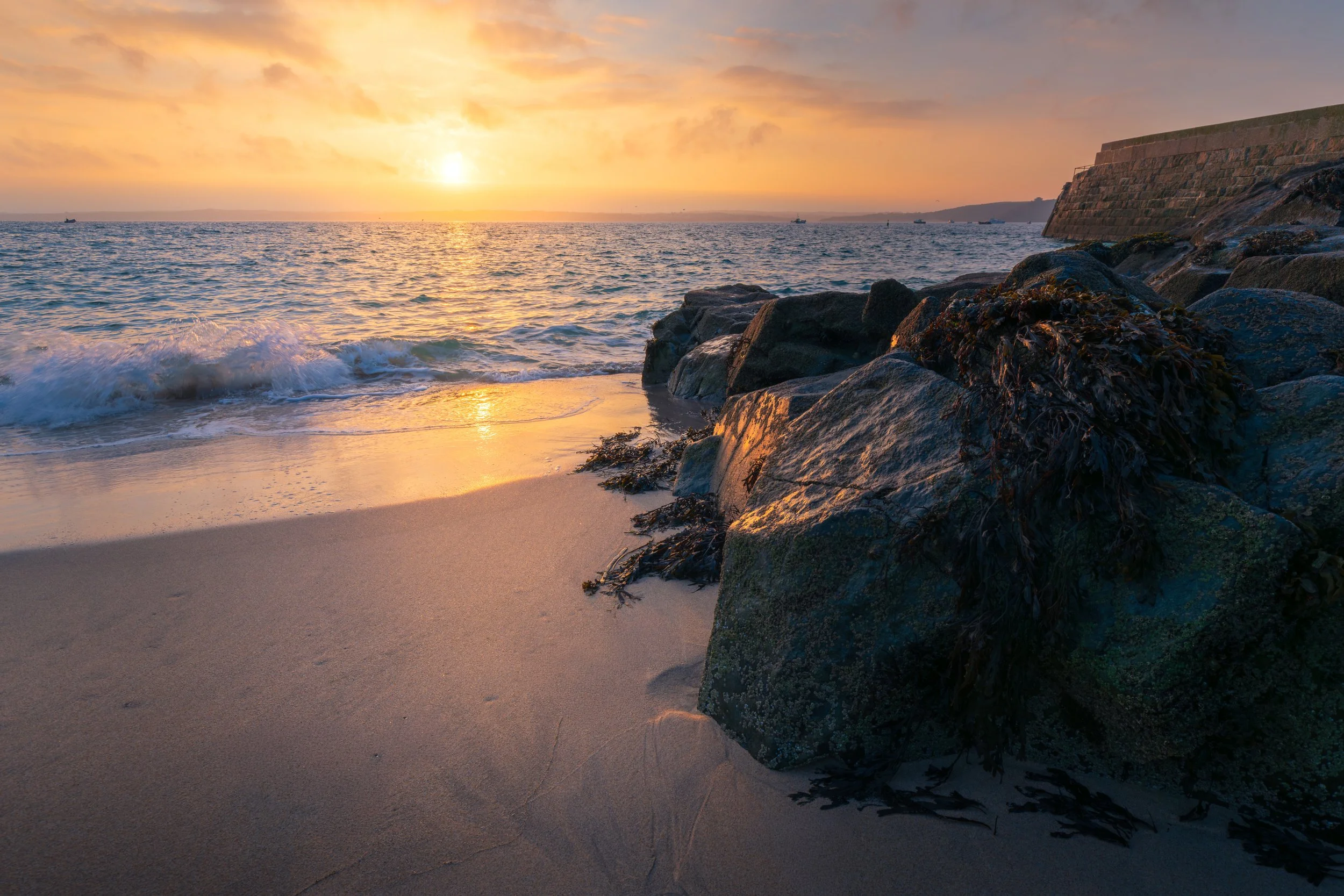 Sunset over the ocean with waves crashing on a sandy beach and rocks in the foreground, with a stone structure on the right side.