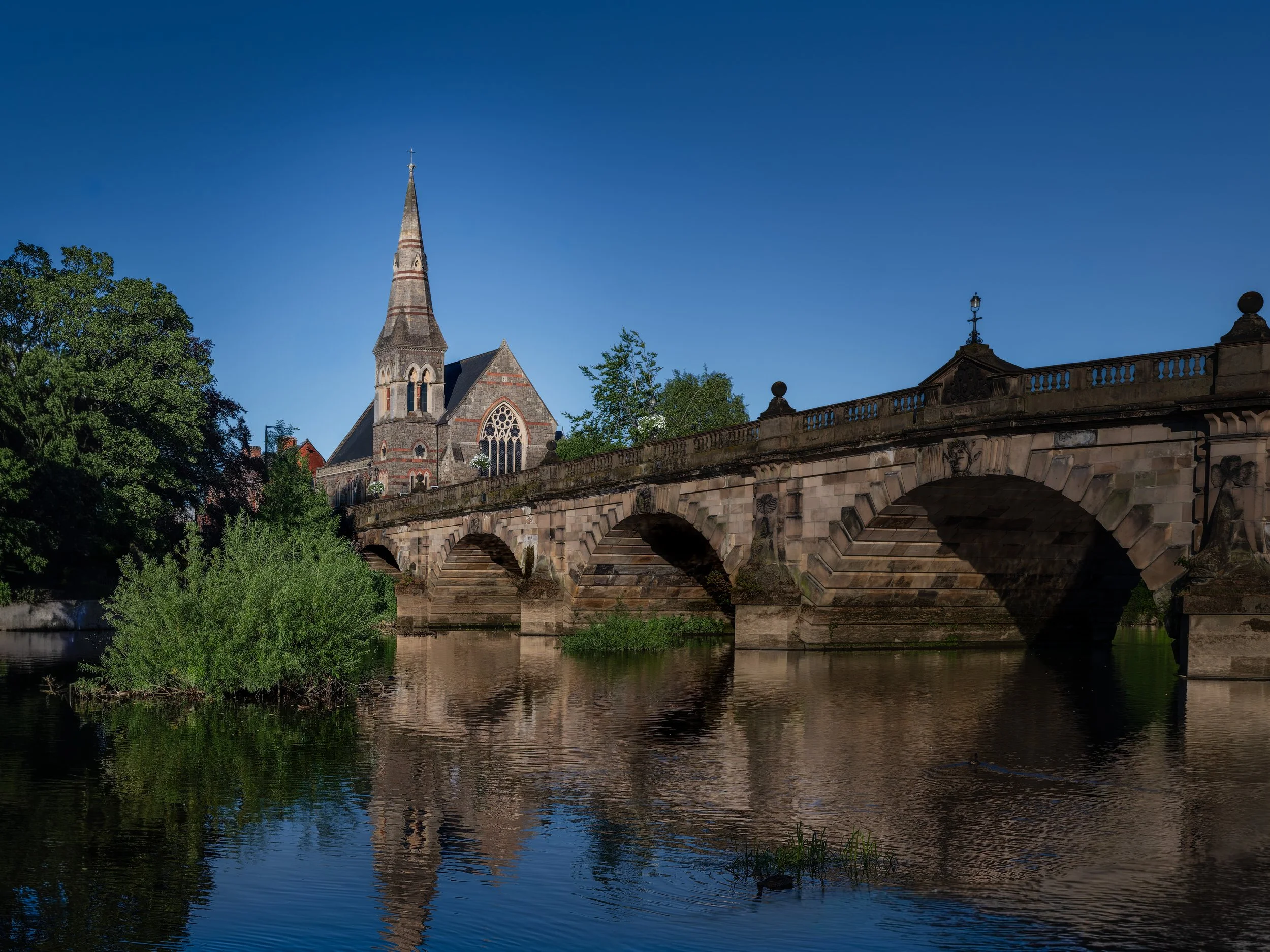 A historic stone bridge over a river with an old church featuring a tall spire in the background, surrounded by trees under a clear blue sky.