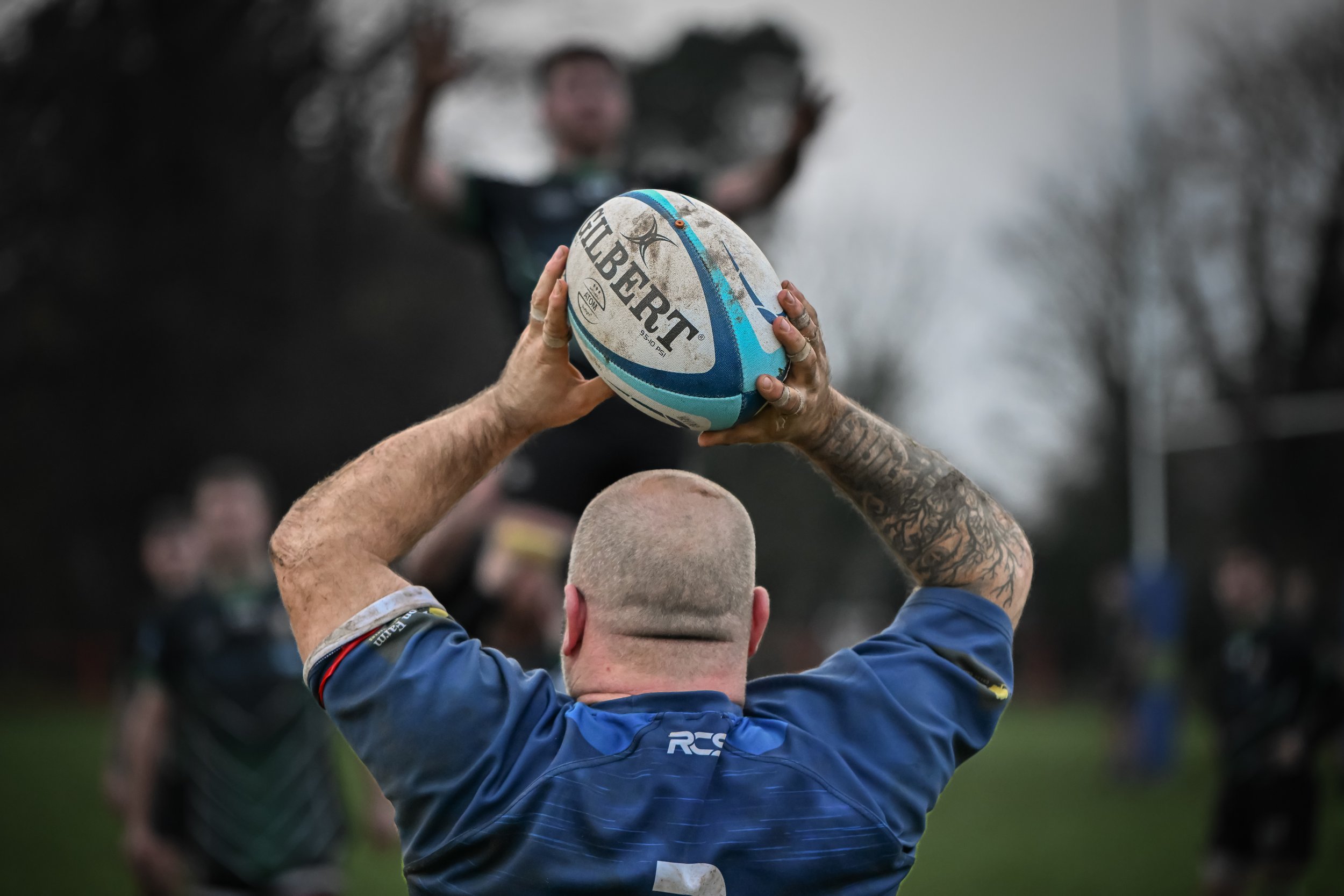 A rugby player with a shaved head and tattoos on his arms is preparing to throw a rugby ball with both hands. The background shows blurred trees and people, including a person in focus standing on a raised area.