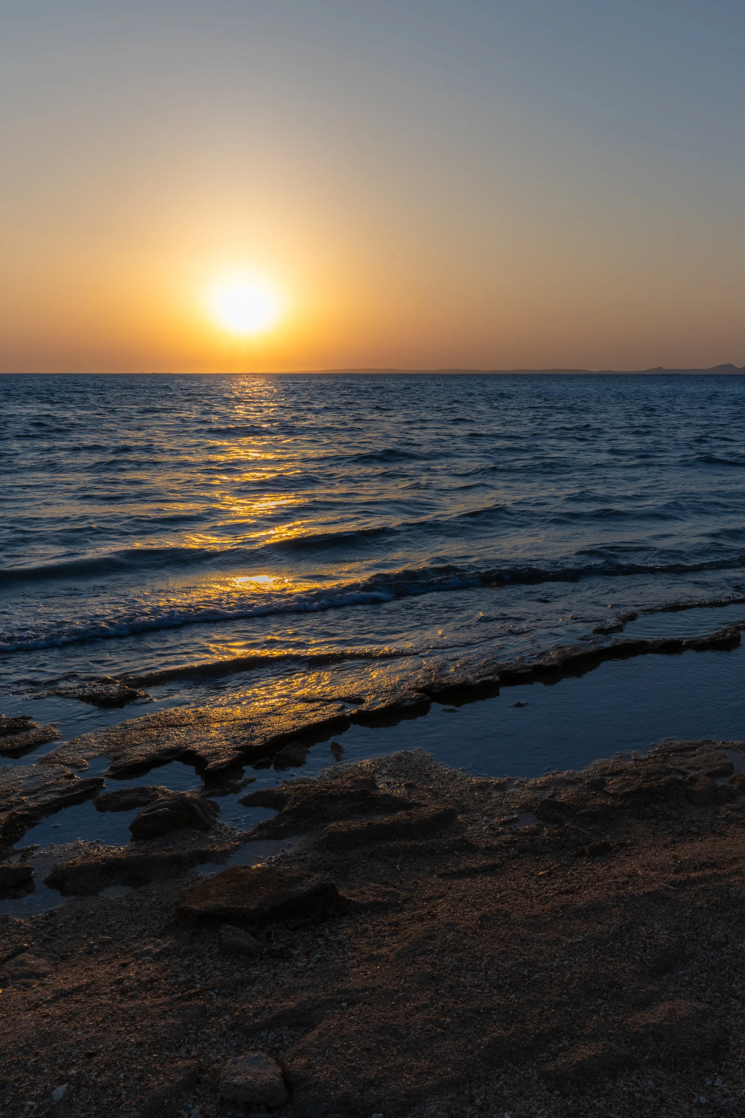 Sunset over the ocean with reflections on the water and rocky beach in the foreground.