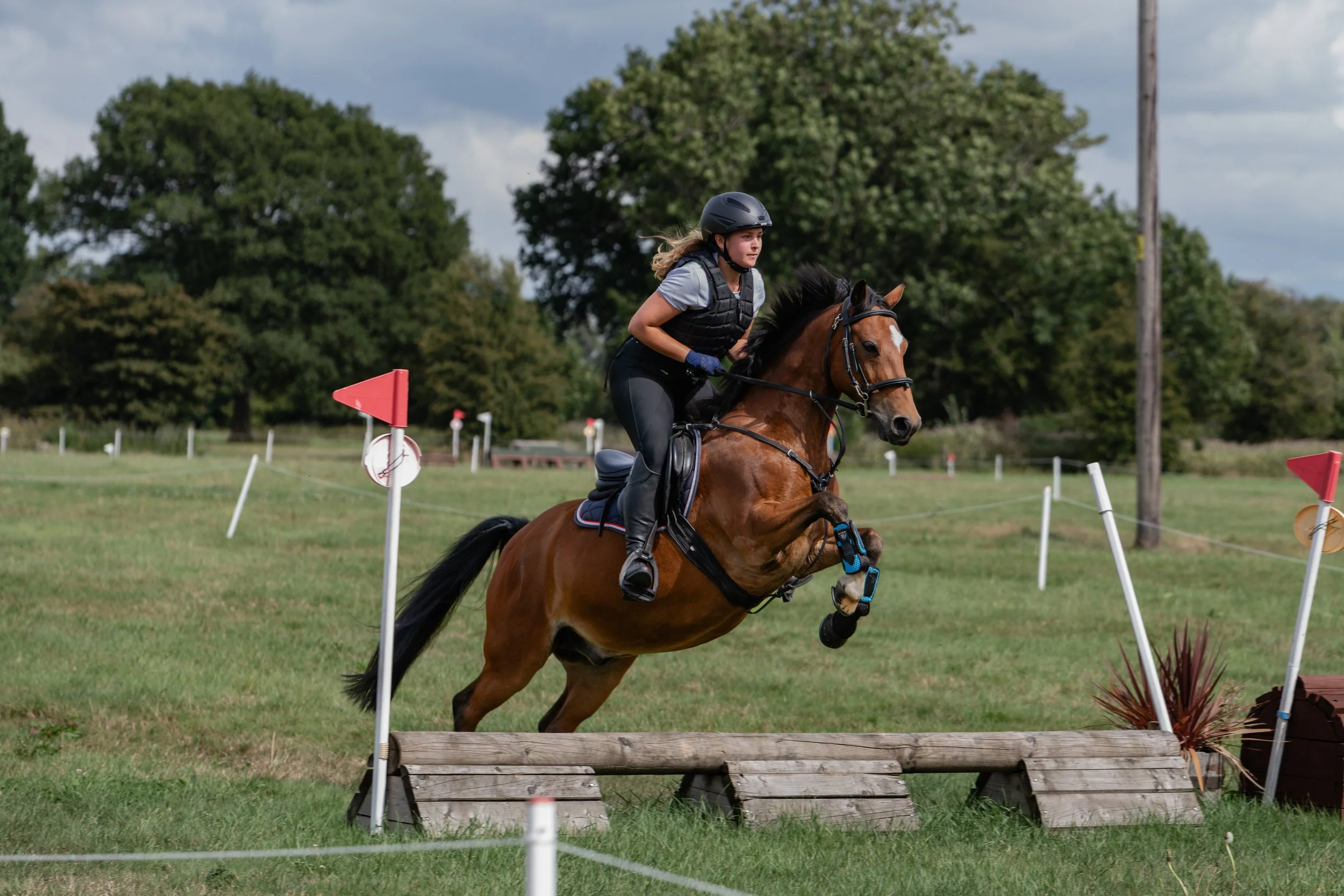 A woman riding a horse jumps over an obstacle during an equestrian event outdoors with trees and a cloudy sky in the background.