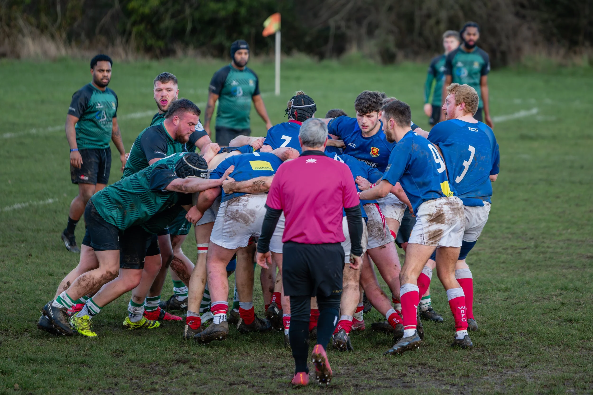 Rugby players engaging in a tackle during a match on a muddy field, with referees and other players observing.