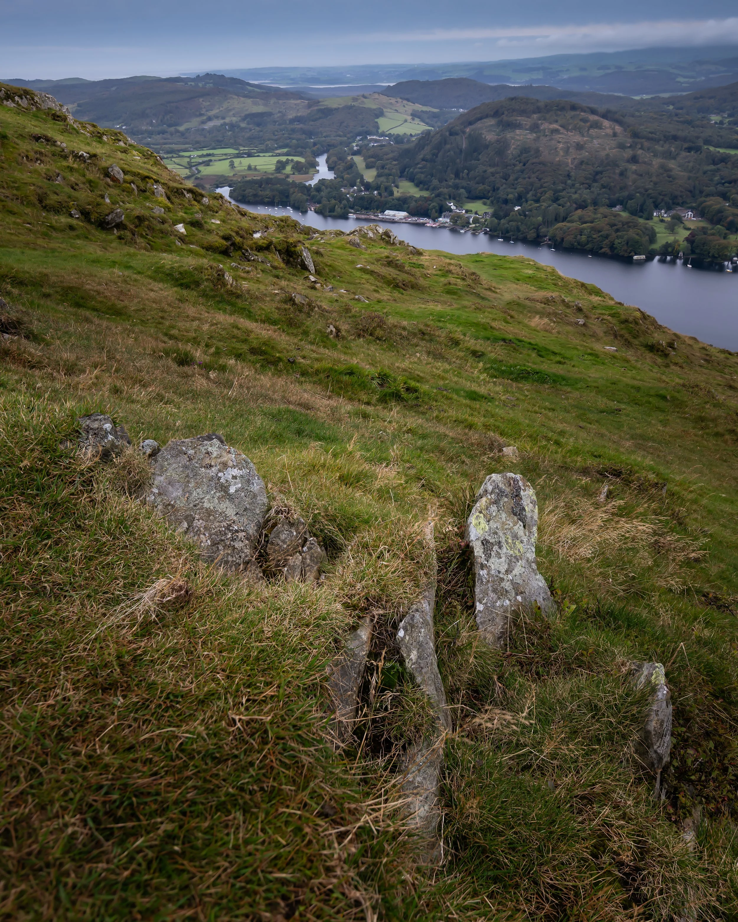 View of a lush green hillside with rocks, overlooking a river and distant hills under a cloudy sky.
