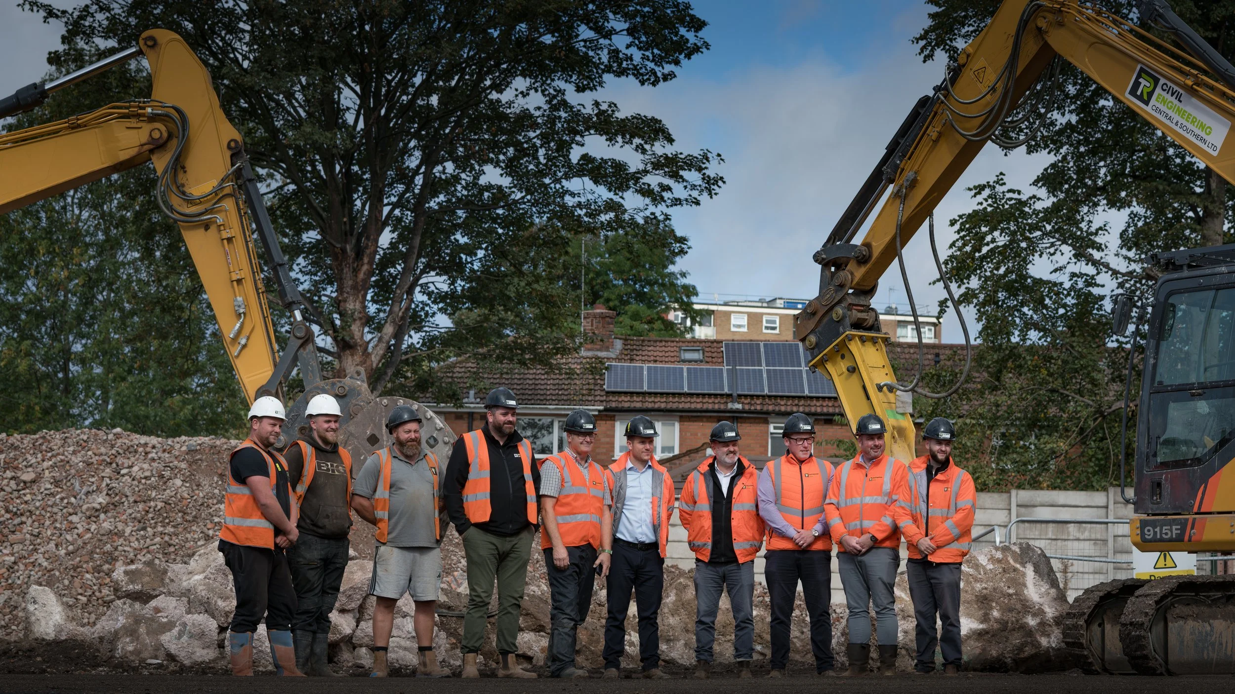 Group of construction workers and engineers standing in front of a construction site with excavators and large rocks, wearing safety helmets and orange safety vests.