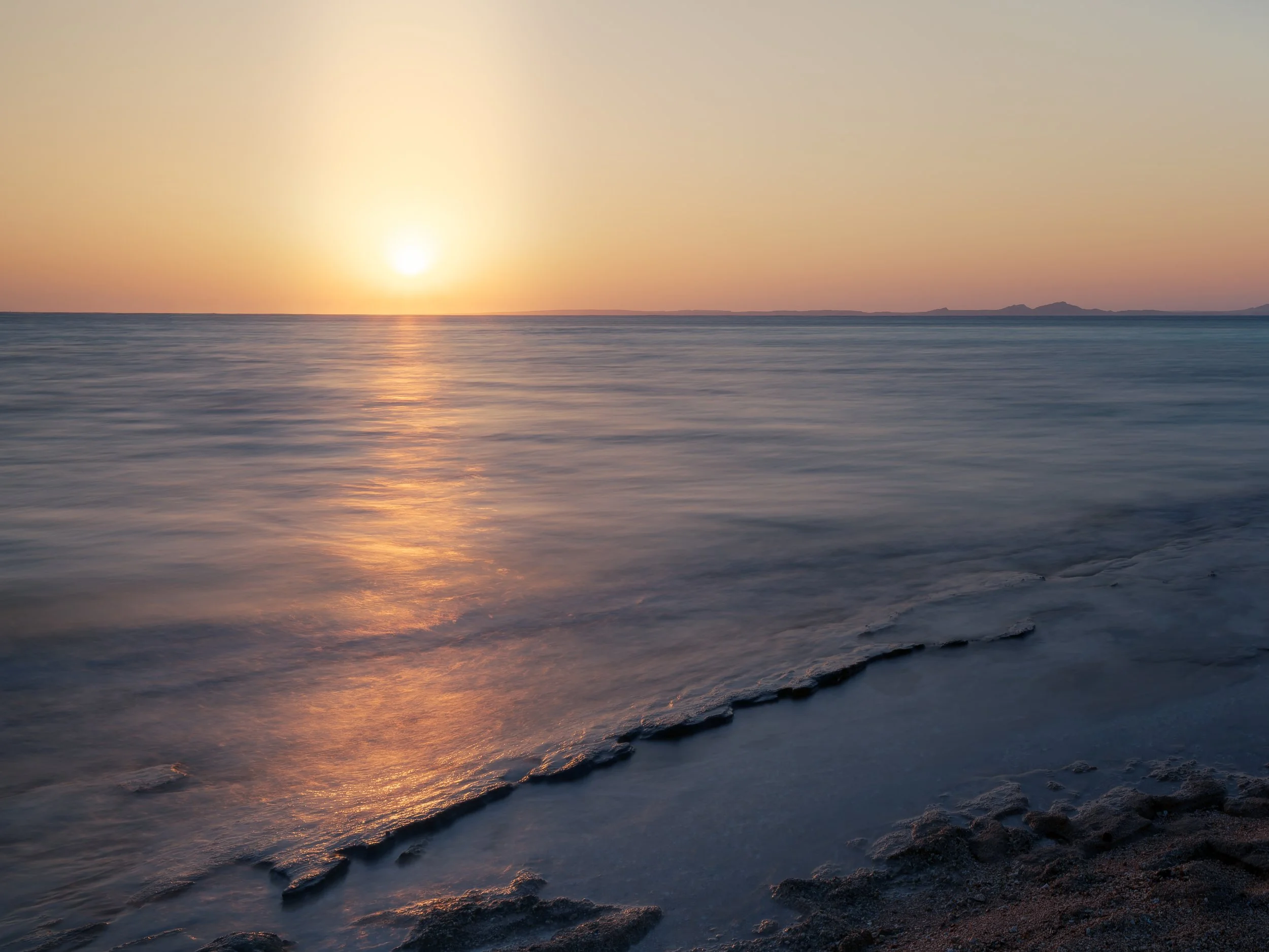 Sunset over calm ocean water with gentle waves and rocky shoreline in the foreground