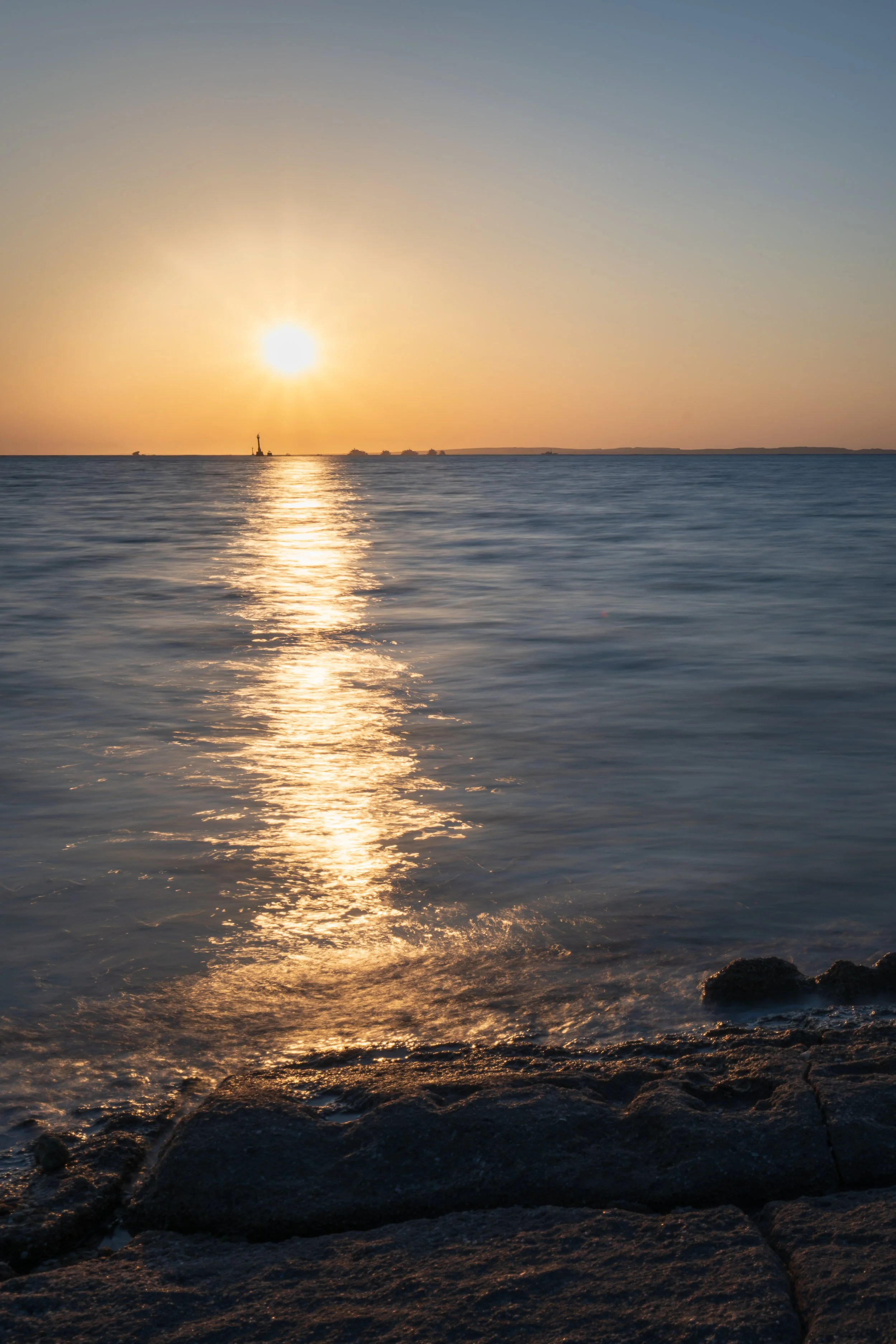 Sunset over the ocean with a reflection on the water, rocks in the foreground, and a distant shoreline with ships.