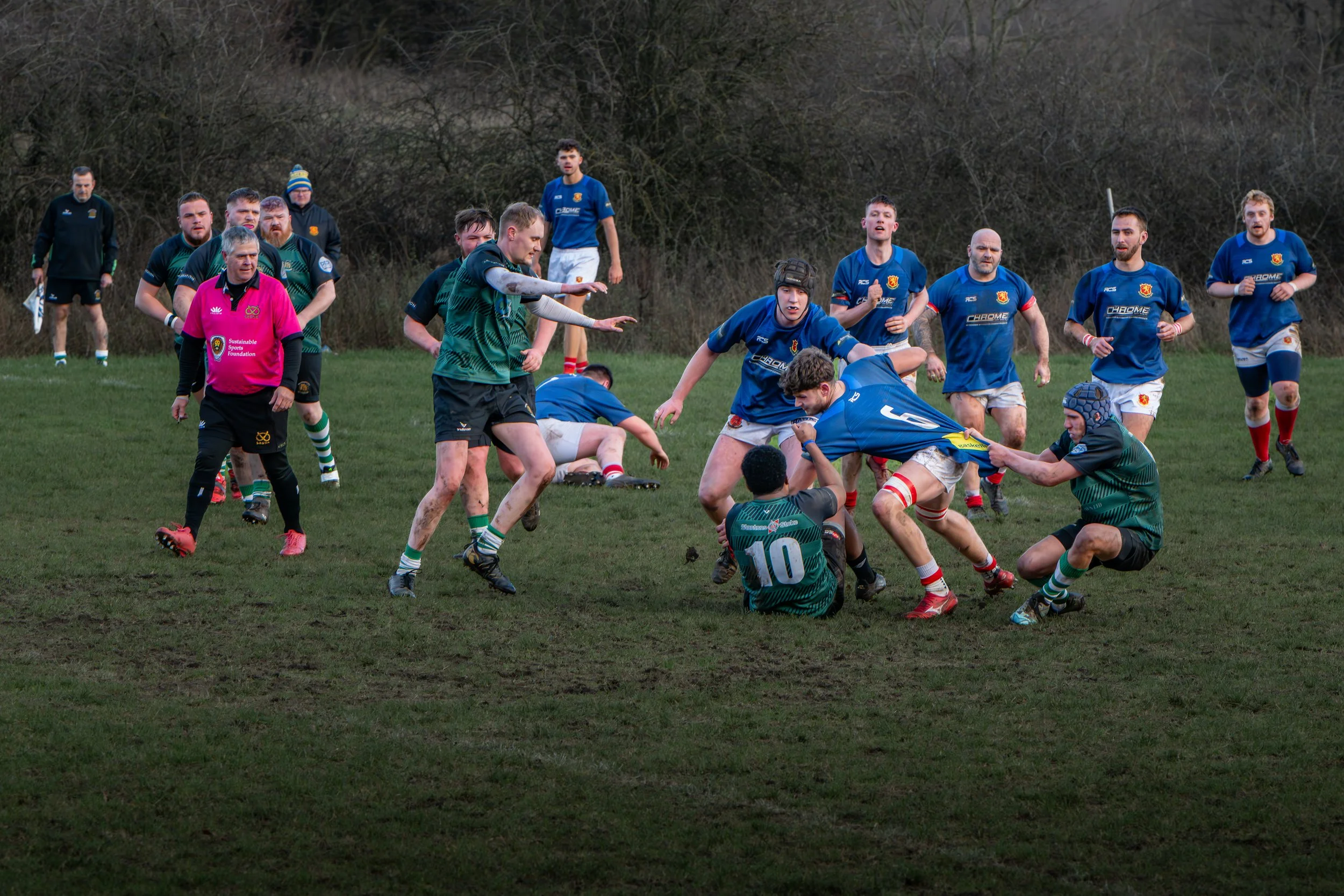 A rugby match in progress with players from two teams, one in green and black uniforms and the other in blue uniforms, engaged in a tackle on a muddy field. Several players are watching and running in the background.
