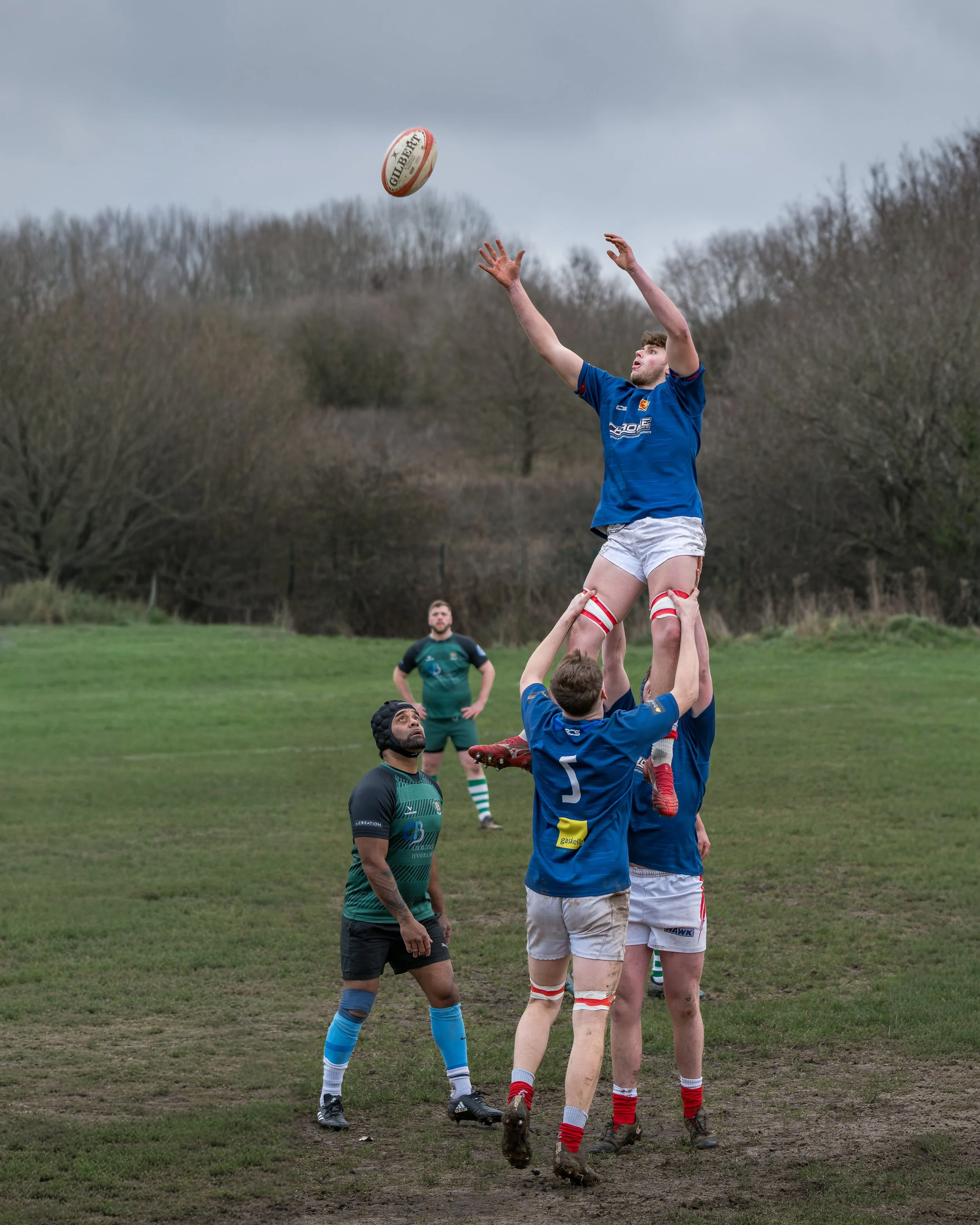 Rugby players contesting lineout, with one player catching the ball while lifted by teammates, on a grassy field with trees and cloudy sky in the background.
