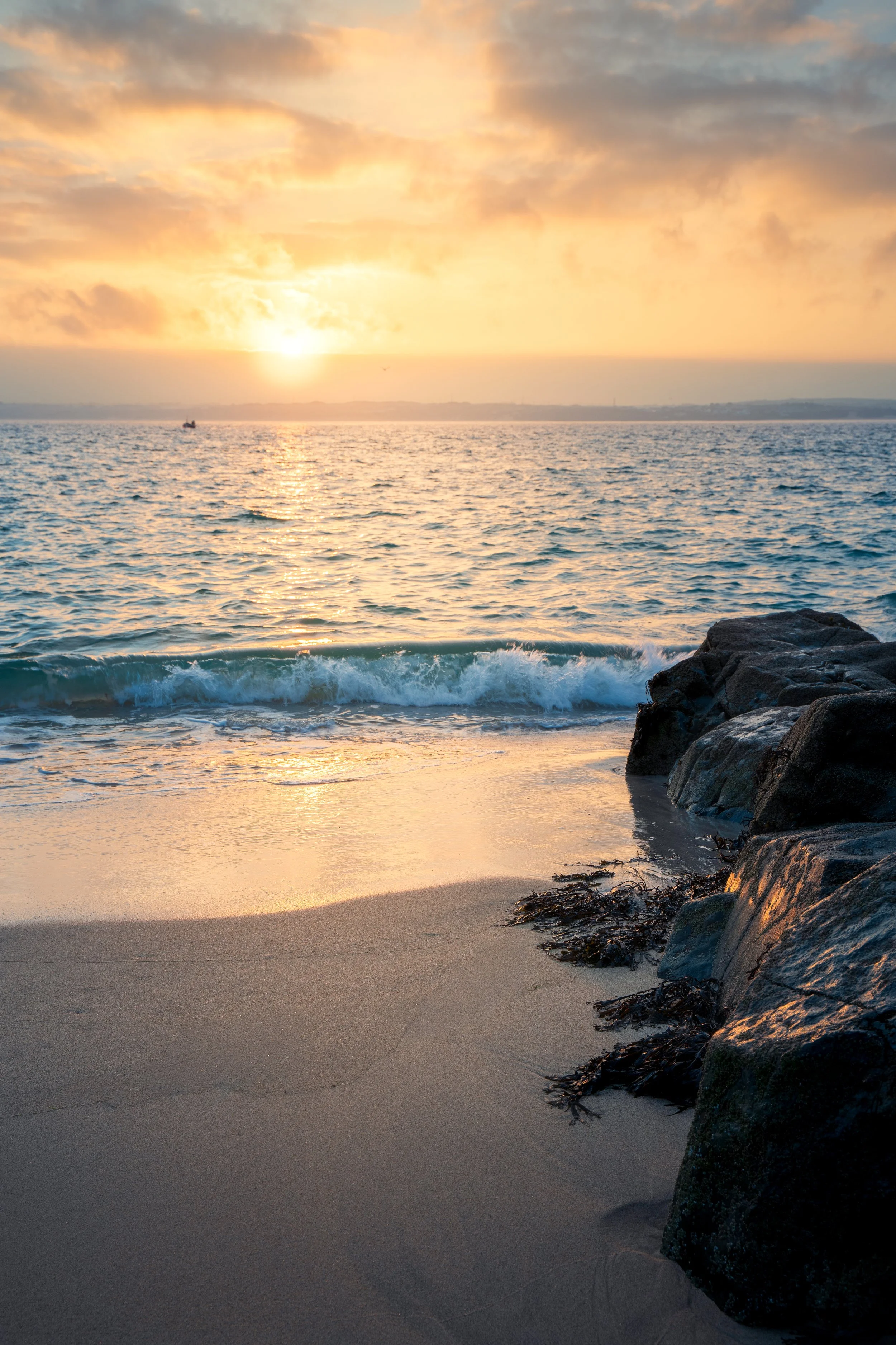Sunset over the ocean with waves near a sandy beach and rocks on the right side.