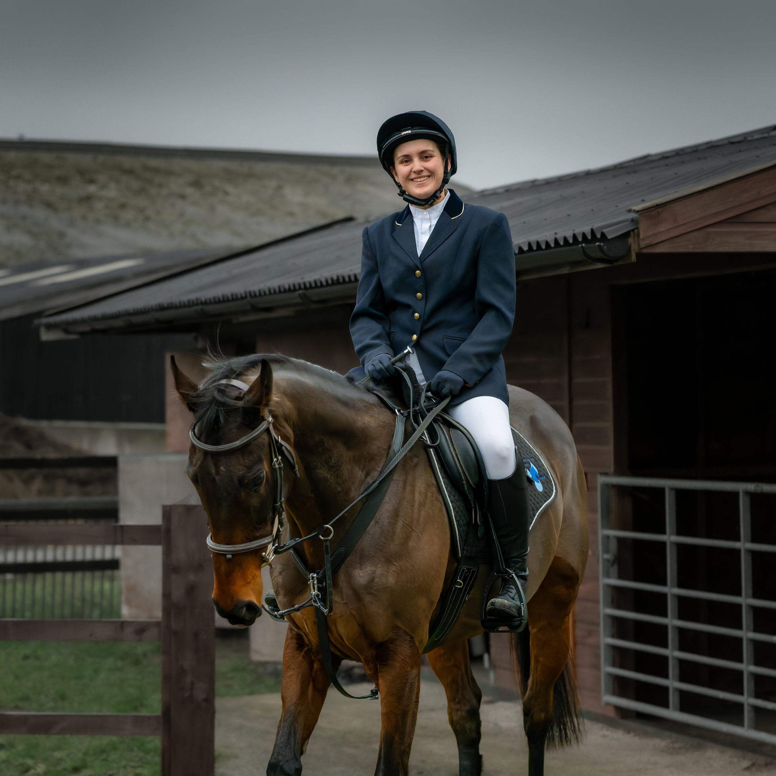 A young woman in equestrian riding attire on a brown horse at a stable.