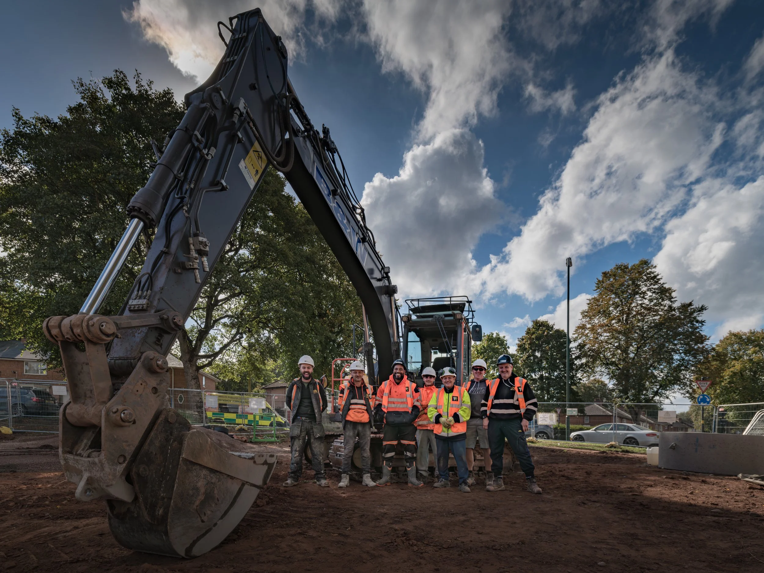 Construction workers standing in front of a large hydraulic excavator on a construction site outdoors with trees and a partly cloudy sky.